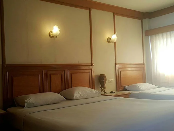 Two beds in a hotel room with wooden headboards, two wall-mounted lamps, a small nightstand with a table lamp, and a window with curtains.