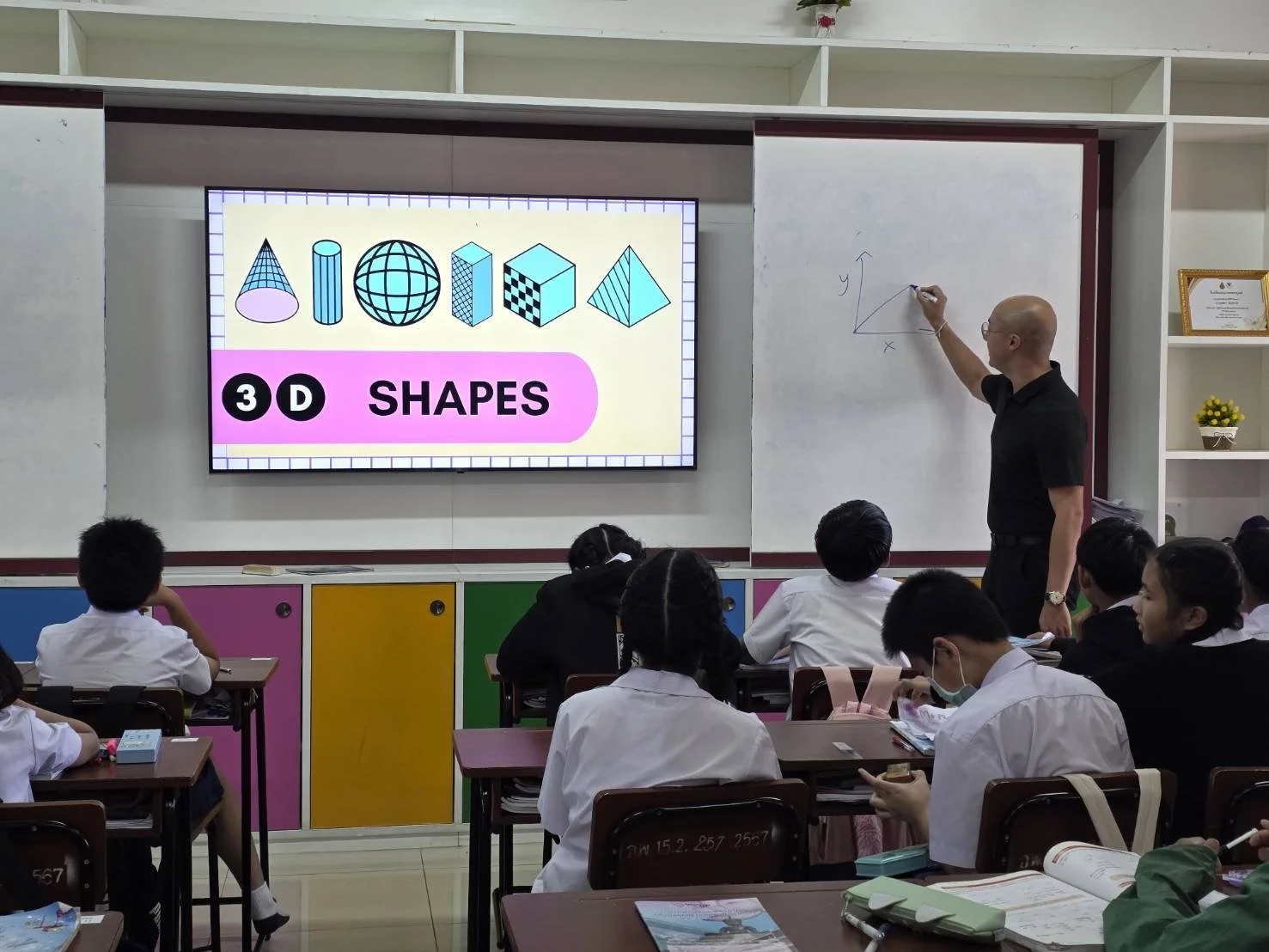 Classroom scene with teacher from Kids English Thailand at whiteboard drawing a graph, students seated at desks, one student in mask, large screen displaying colorful 3D shapes and the text '3D SHAPES'.