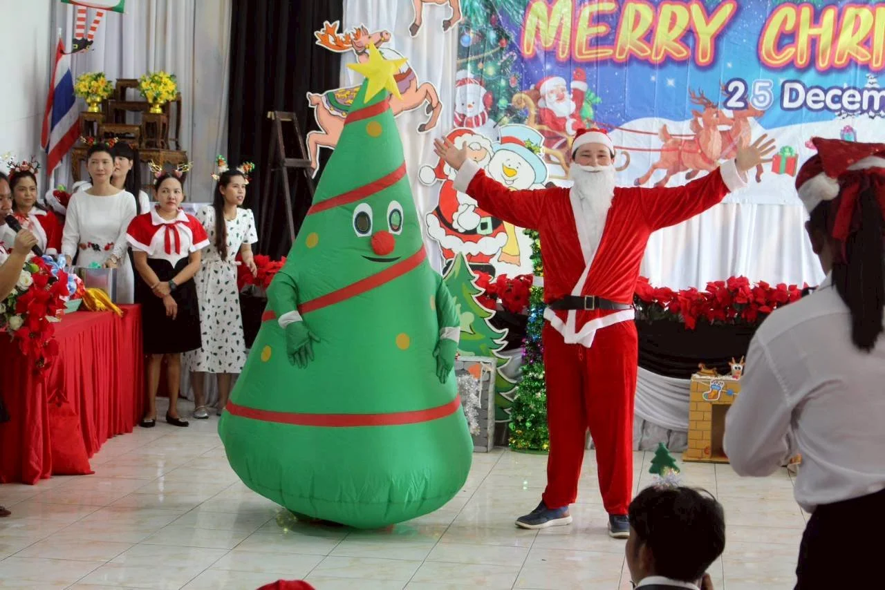 Kids English Thailand teachers celebrating Christmas with a person dressed as Santa Claus, a Christmas tree inflatable with a face, and a group of women in holiday costumes. There is a festive backdrop with Christmas characters and decorations.