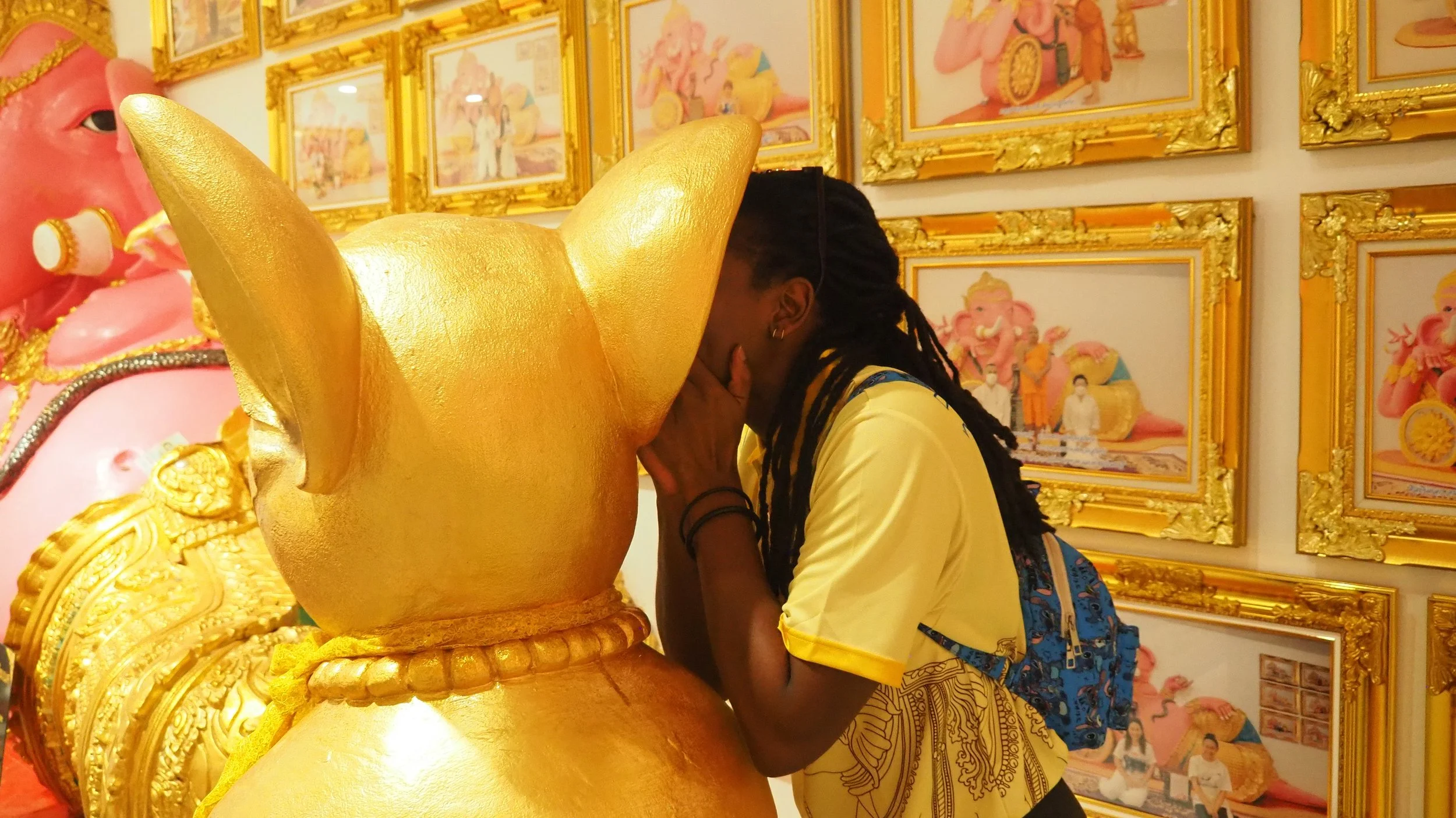 KET Teacher with long braids, wearing a yellow shirt, faces a large golden statue with a rounded head and big ears, and has hands clasped in prayer or reverence. 