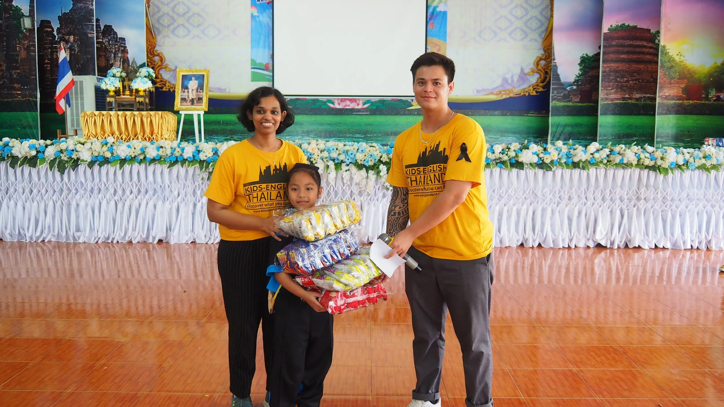 Two Kids English Thailand teachers  standing on a stage during an event, with a woman, a man, and a girl holding packages of snacks or supplies, wearing yellow t-shirts with event logos.