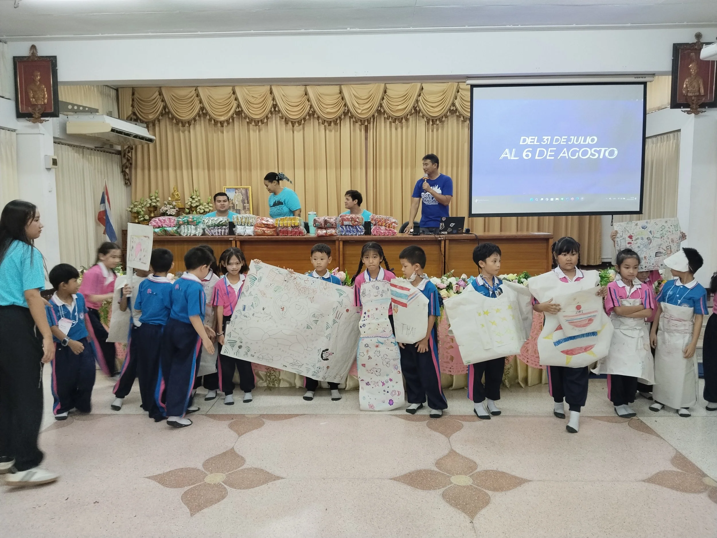 School children in uniforms holding decorated paper bags or posters standing in front of a stage with a large screen displaying a message, and with Kids English Thailand teachers at a table behind them in a decorated hall.