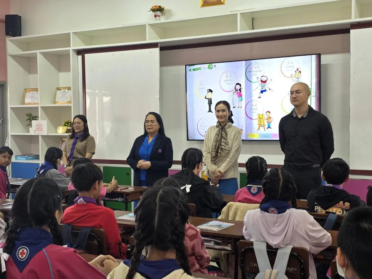 Students seated at desks in a classroom listening to four teachers from Kids English Thailand standing in front of a large digital screen displaying colorful cartoon characters and text.