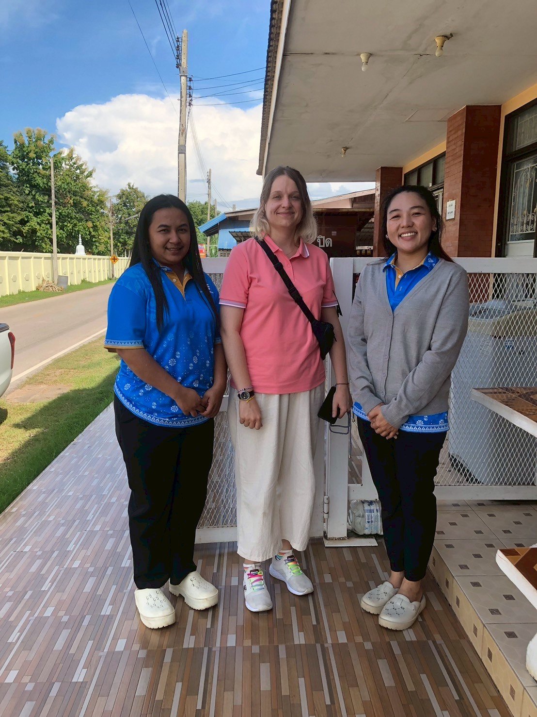 Three women from Kids English Thailand standing outdoors on a wooden porch, smiling at the camera with a street, trees, and a building in the background.