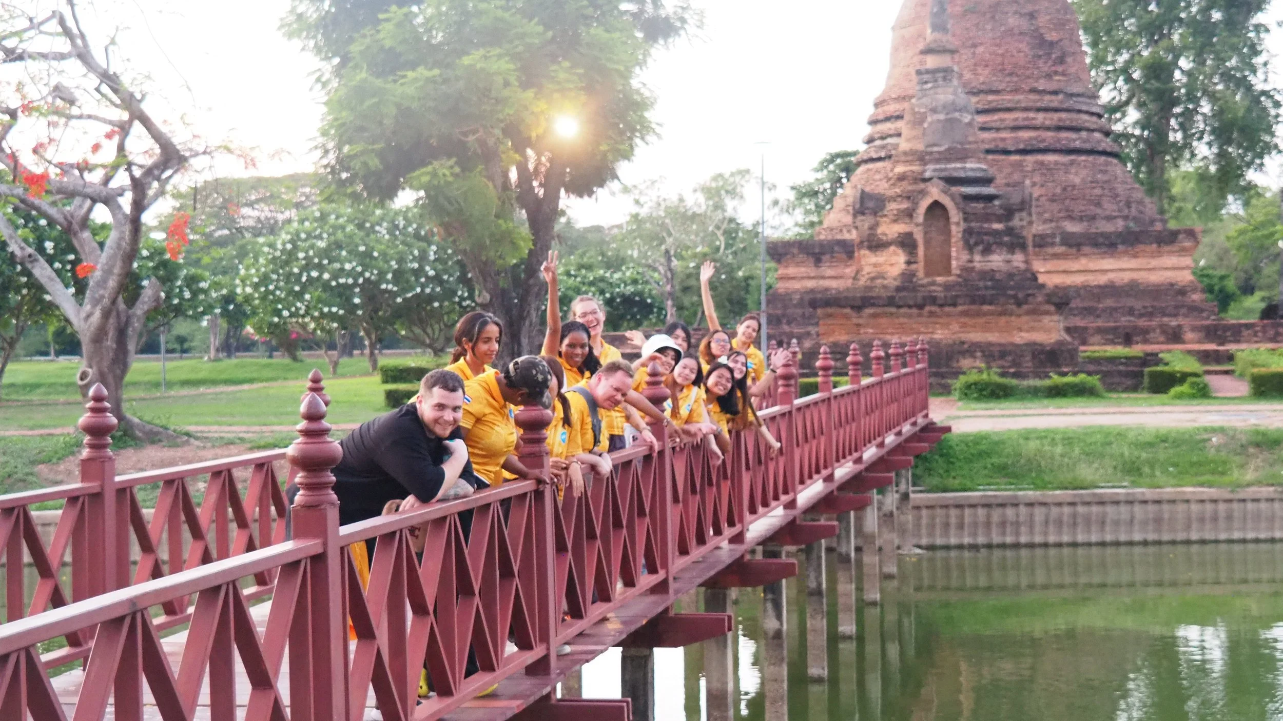 Group of Kids English Thailand teachers on a red bridge at a historical site with a brick stupa in the background, surrounded by trees and greenery in the evening with sunlight.