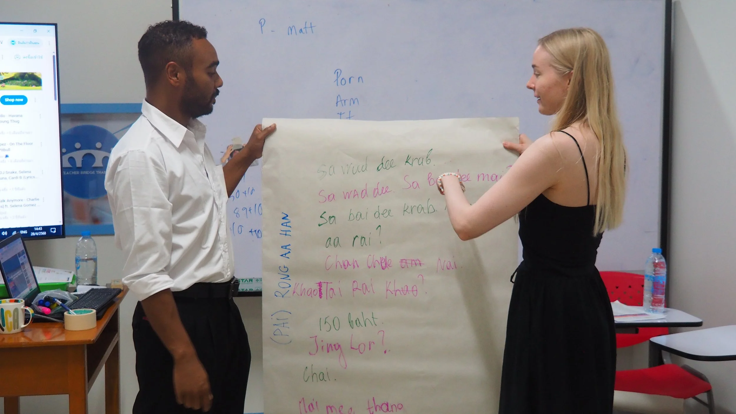 Two Kids English Thailand teachers, are holding a large sheet of paper with handwritten text in front of a whiteboard in a classroom. 