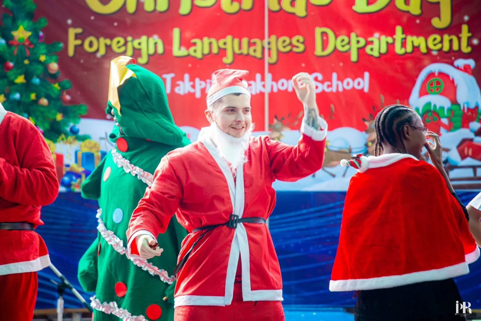 Kids English Thailand teacher dressed as Santa Claus waving at a Christmas event, with a decorated Christmas tree and holiday backdrop in the background.