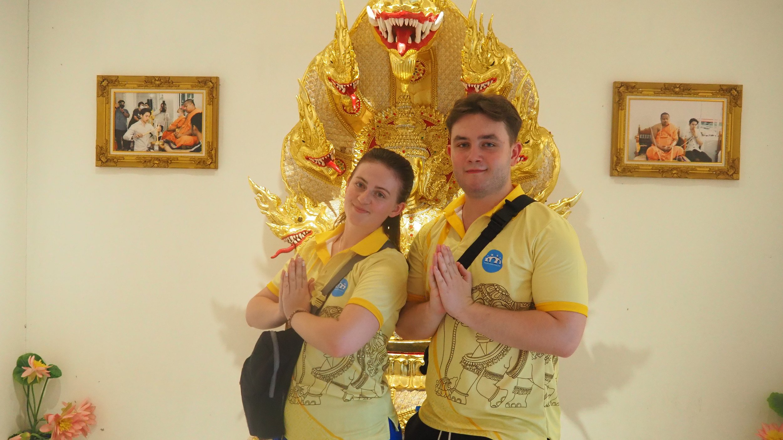 A  couple of Kids English Thailand teachers in yellow matching shirts with traditional elephant and mythical creature designs stand in prayer with hands pressed together, in front of ornate gold and red dragon decor, inside a temple.