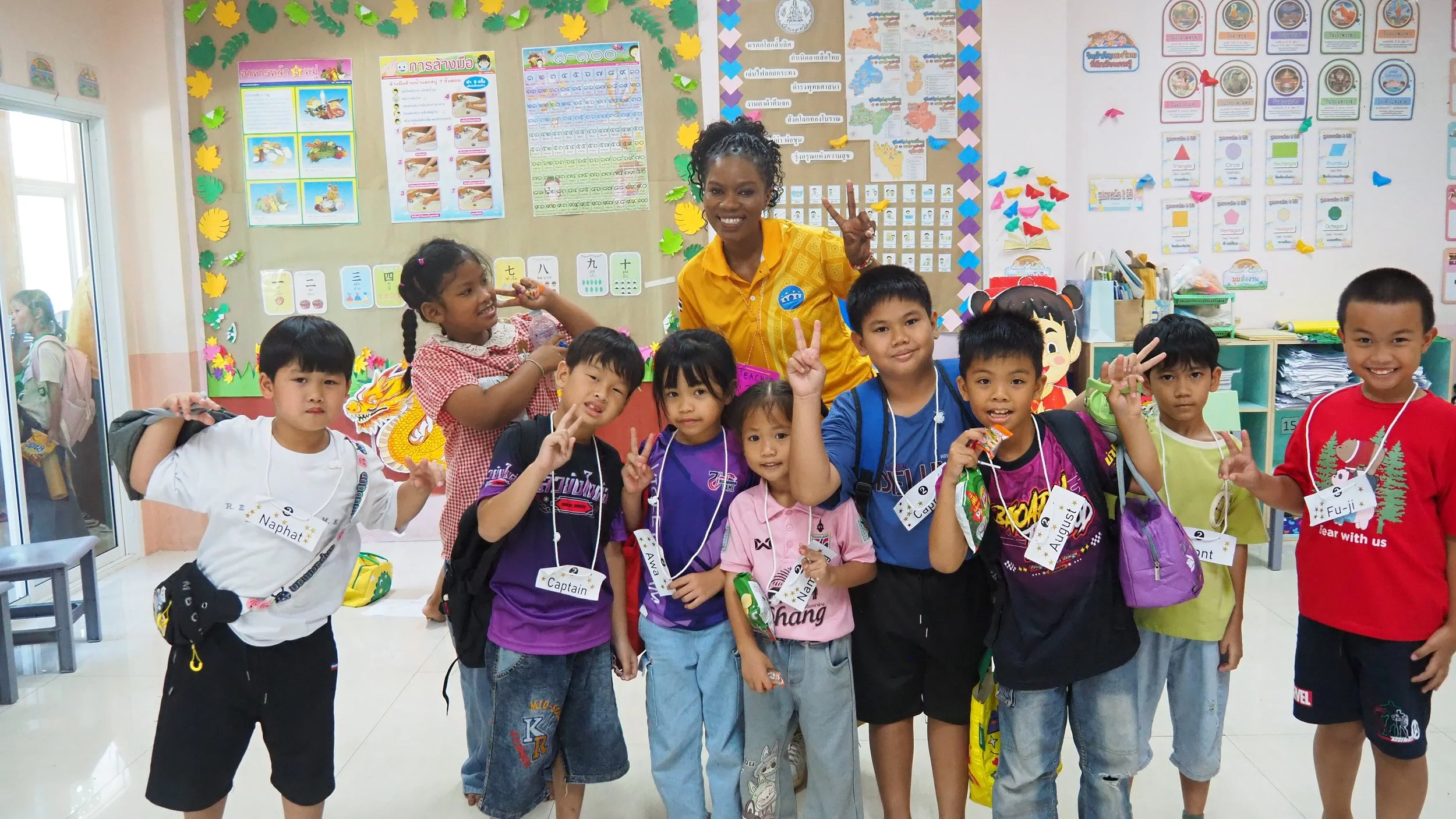 Group of young KET children and their Kids English Thailand teacher in a classroom, smiling and holding up peace signs, with colorful educational posters and decorations behind them.