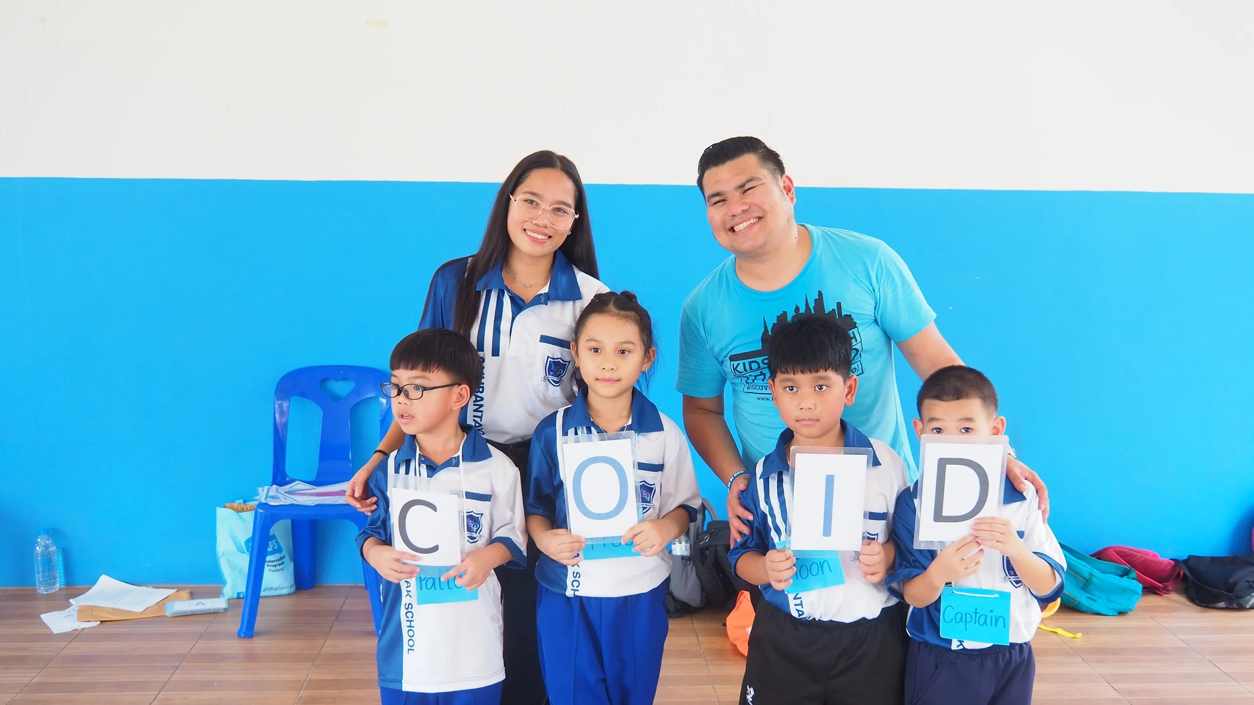 Four young children in school uniforms holding signs that spell out 'COID', standing in front of a blue wall with two Kids English Thailand teachers standing behind them, all smiling.