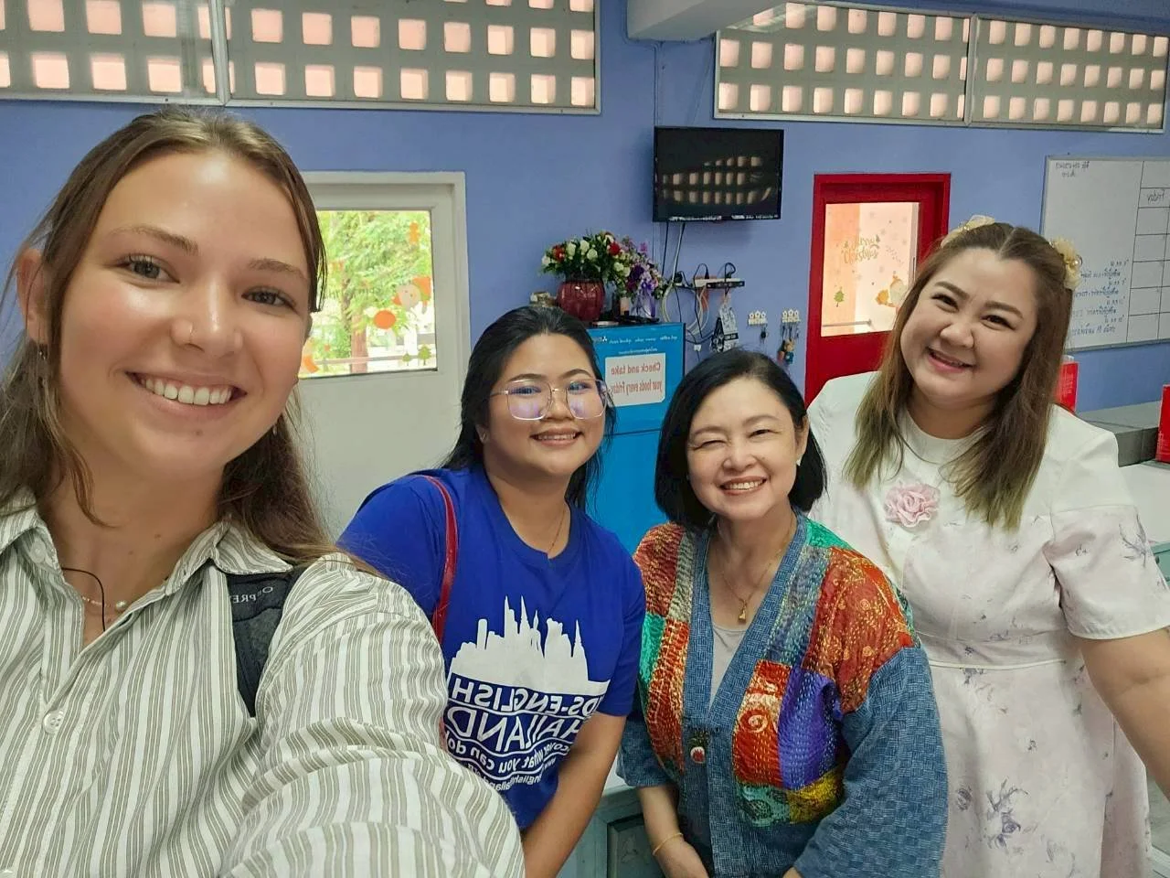 Four women from Kids English Thailand smiling together in an indoor setting, with a blue wall, window, TV, flowers, and whiteboard in the background.