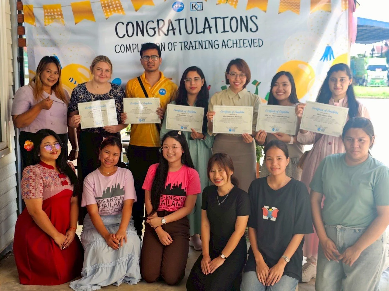 Group of Kids English Thailand teachers, interns and volunteers at a training completion ceremony, holding certificates in front of a decorated backdrop that says 'CONGRATULATIONS' and 'COMPLETION OF TRAINING ACHIEVED'.