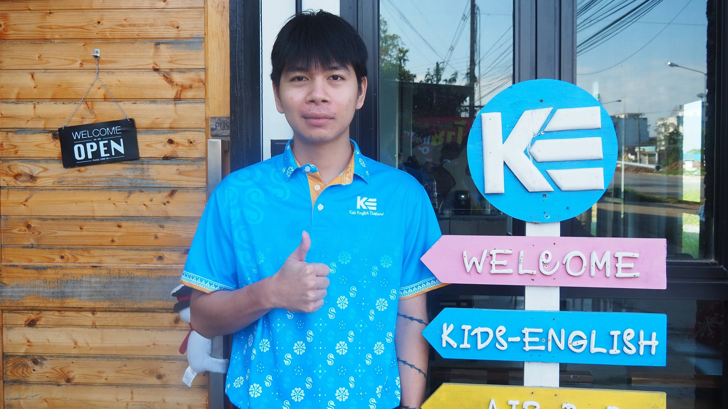 Young man standing in front of a school entrance, wearing a blue shirt with the Kids English Thailand logo, giving a thumbs-up gesture.