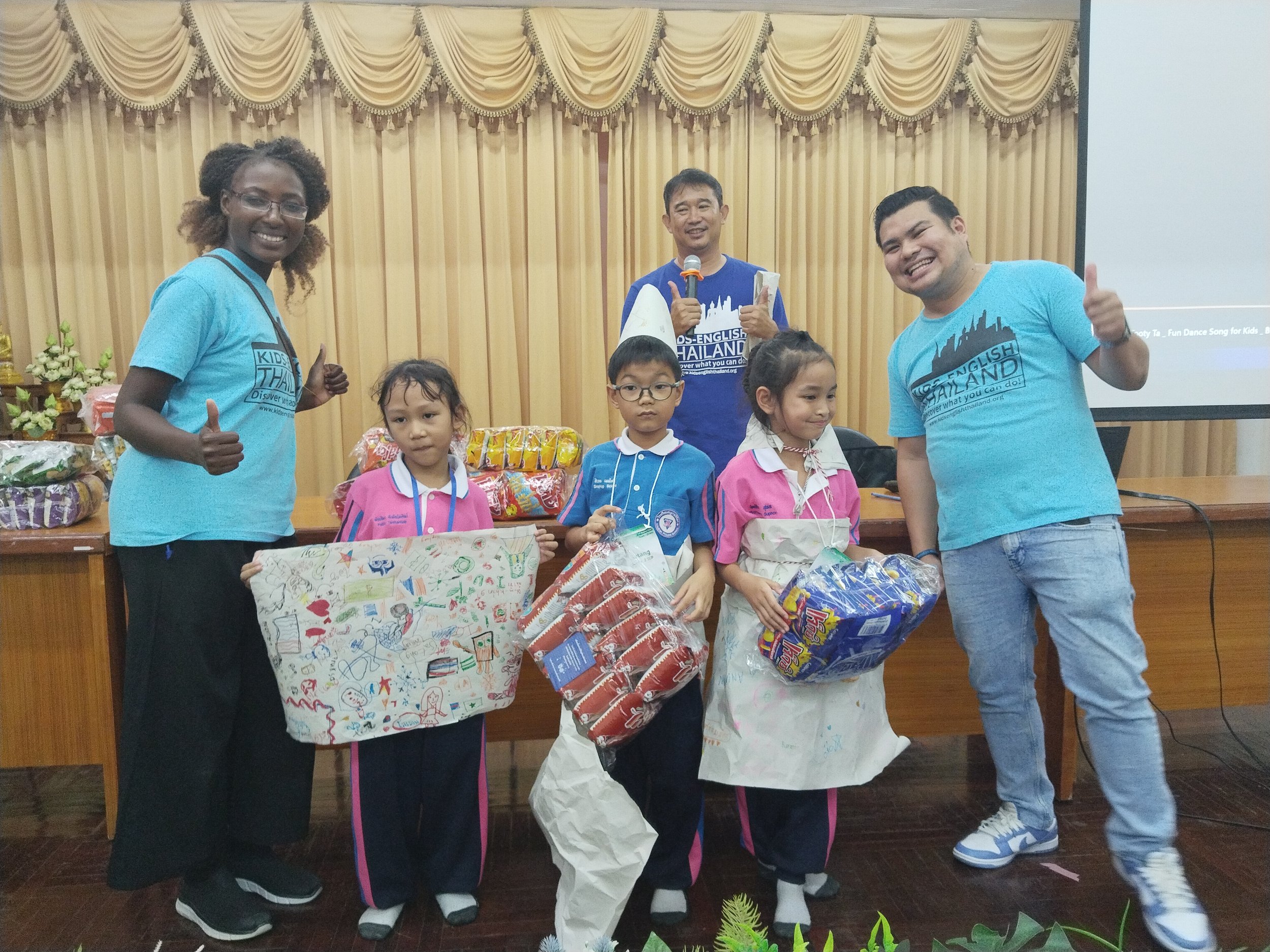 Group photo of children and Kids English Thailand teachers at an award ceremony, with children holding gifts and wearing medals, in front of golden curtains and a table with more gifts.