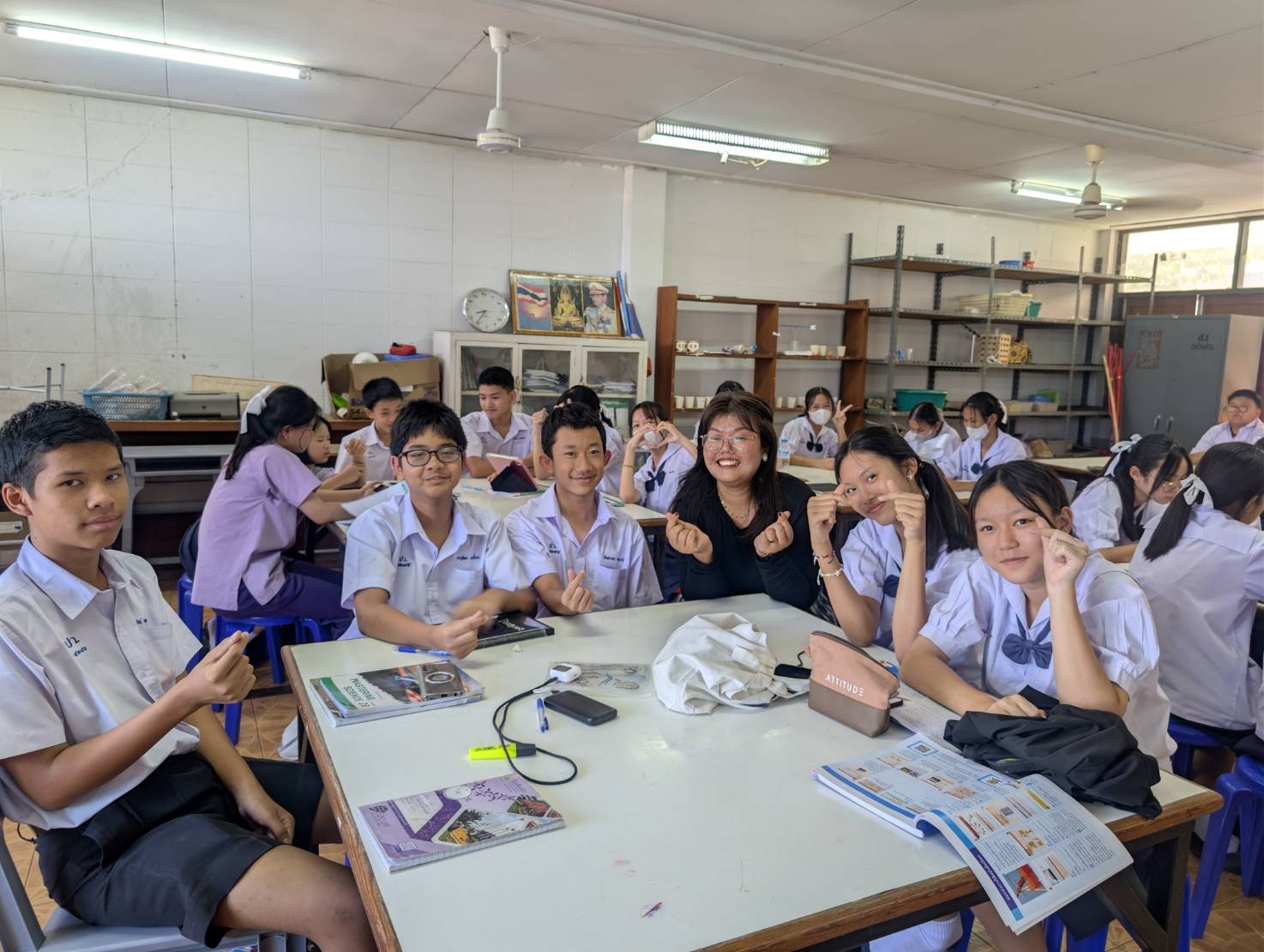 A classroom full of students in school uniforms sitting at desks, with some making finger heart gestures, and a Kids English Thailand teacher smiling at the front. Students are engaged with books and notebooks on the tables.