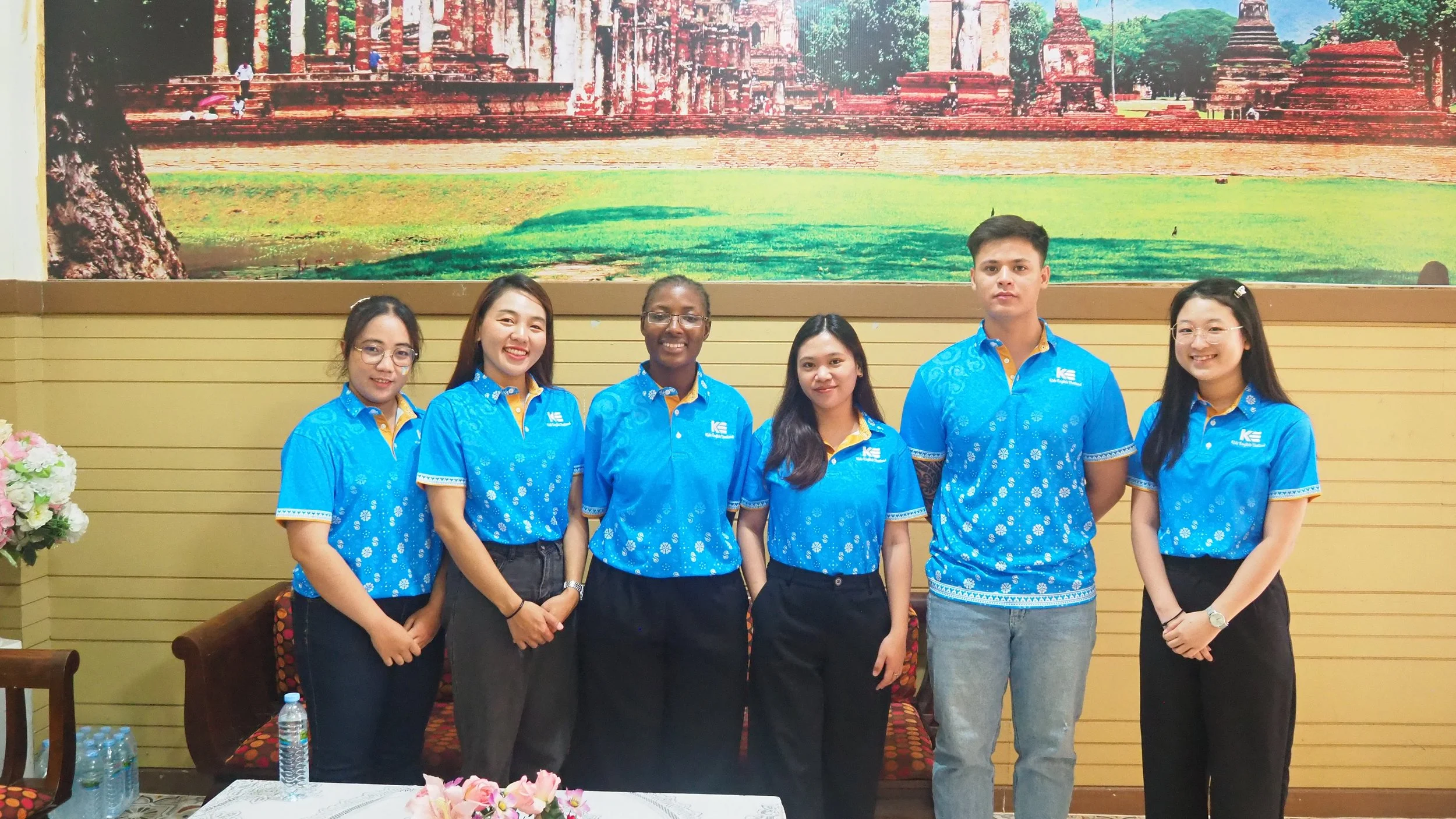 Group of six Kids English Thailand teachers standing indoors in front of a large mural of historical ruins, wearing matching blue KET shirts with a logo, smiling.
