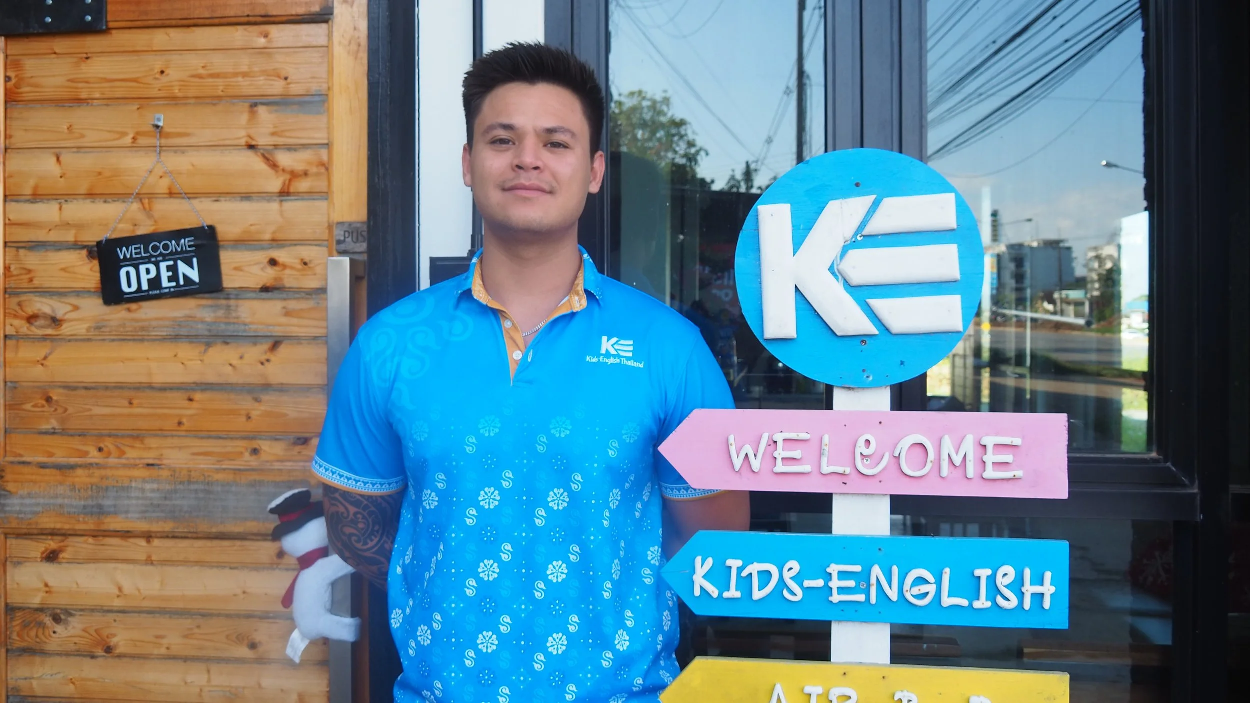 A smiling man with a tattoo on his left arm, wearing a blue patterned shirt, giving a thumbs up while standing outside a building with a large window and colorful signs indicating directions, including 'Welcome,' 'Kids-English,' 'Air BnB,' 'Organic Farm,' and 'Farm House.'