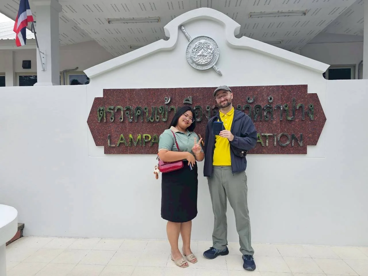 Two people from Kids English Thailand standing in front of a sign that says "Lampang" in Thai and English, smiling and posing for the camera.