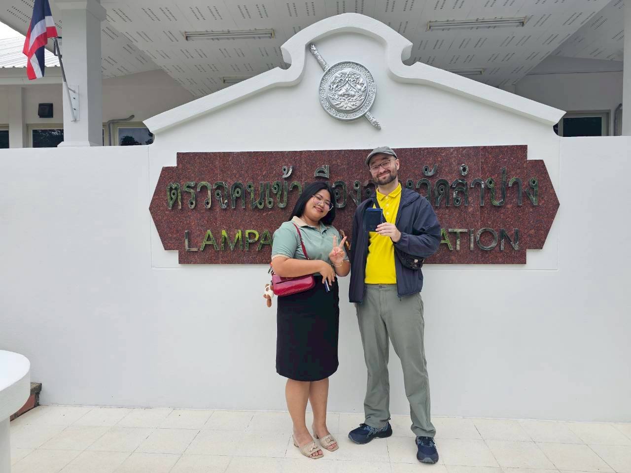Two people from Kids English Thailand standing in front of a sign that says "Lampang" in Thai and English, smiling and posing for the camera.