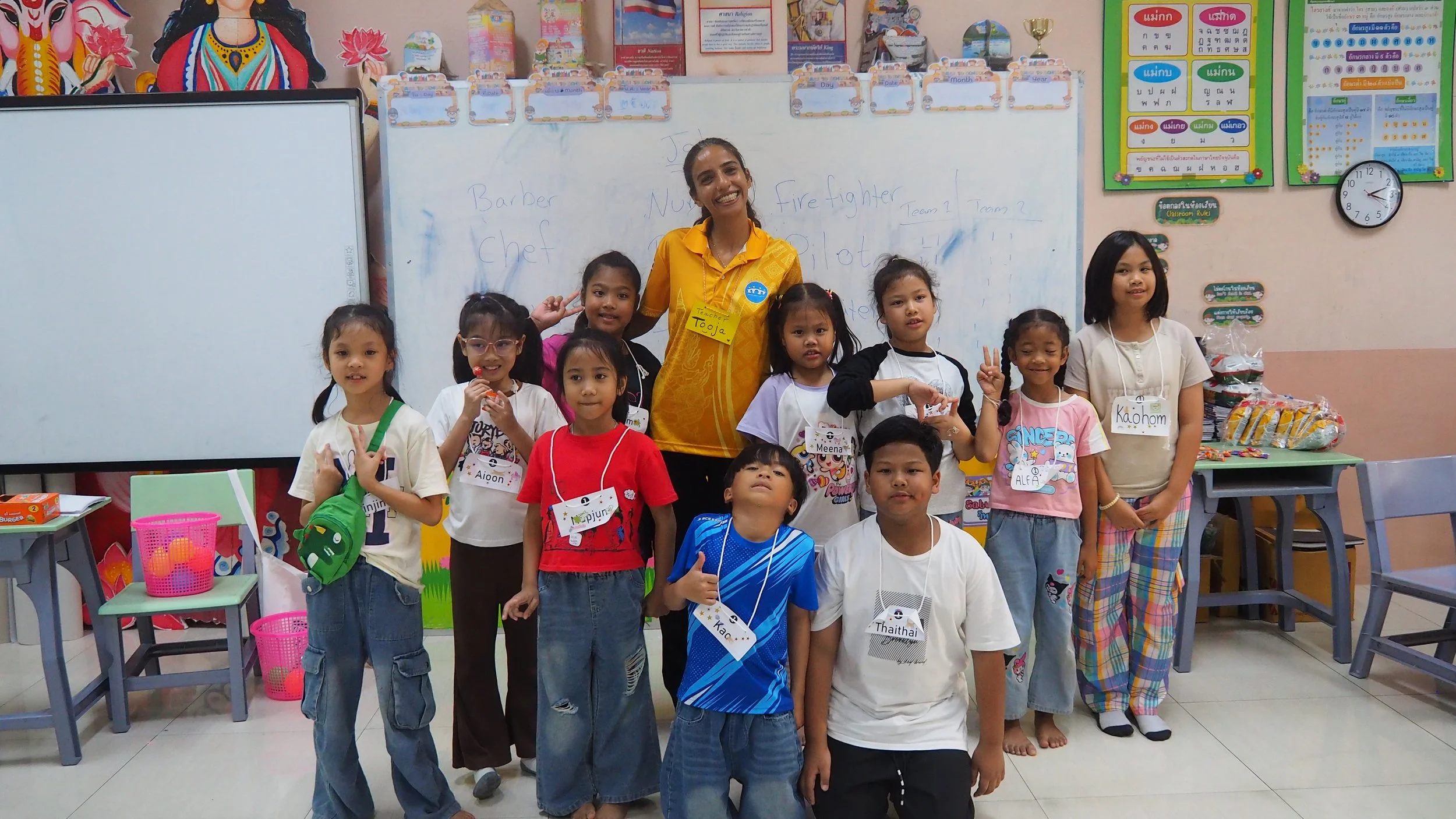 Group of young KET children with a Kids English Thailand teacher in a classroom, some holding up peace signs. The classroom has educational posters, a whiteboard, and a clock on the wall.