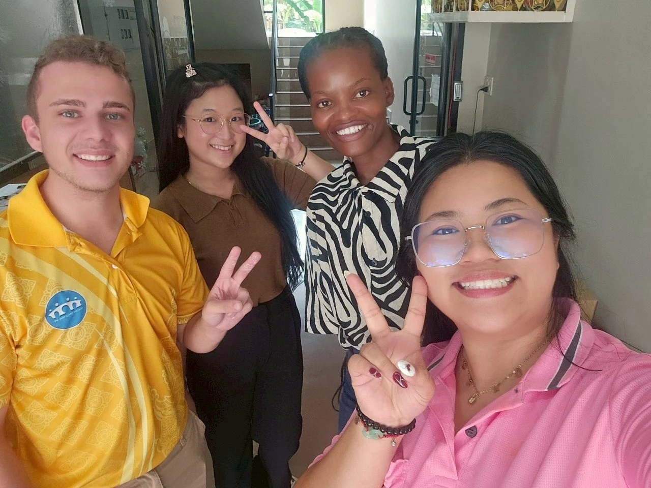 Four young adults from Kids English Thailand smiling and making peace signs in a bright indoor setting, some wearing glasses and casual clothing.