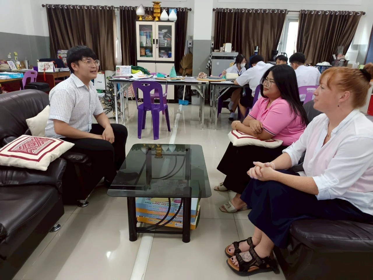 A group of young people from Kids English Thailand are having a conversation in a classroom or office, with a man speaking while three women listen attentively, seated on sofas in front of a glass table.