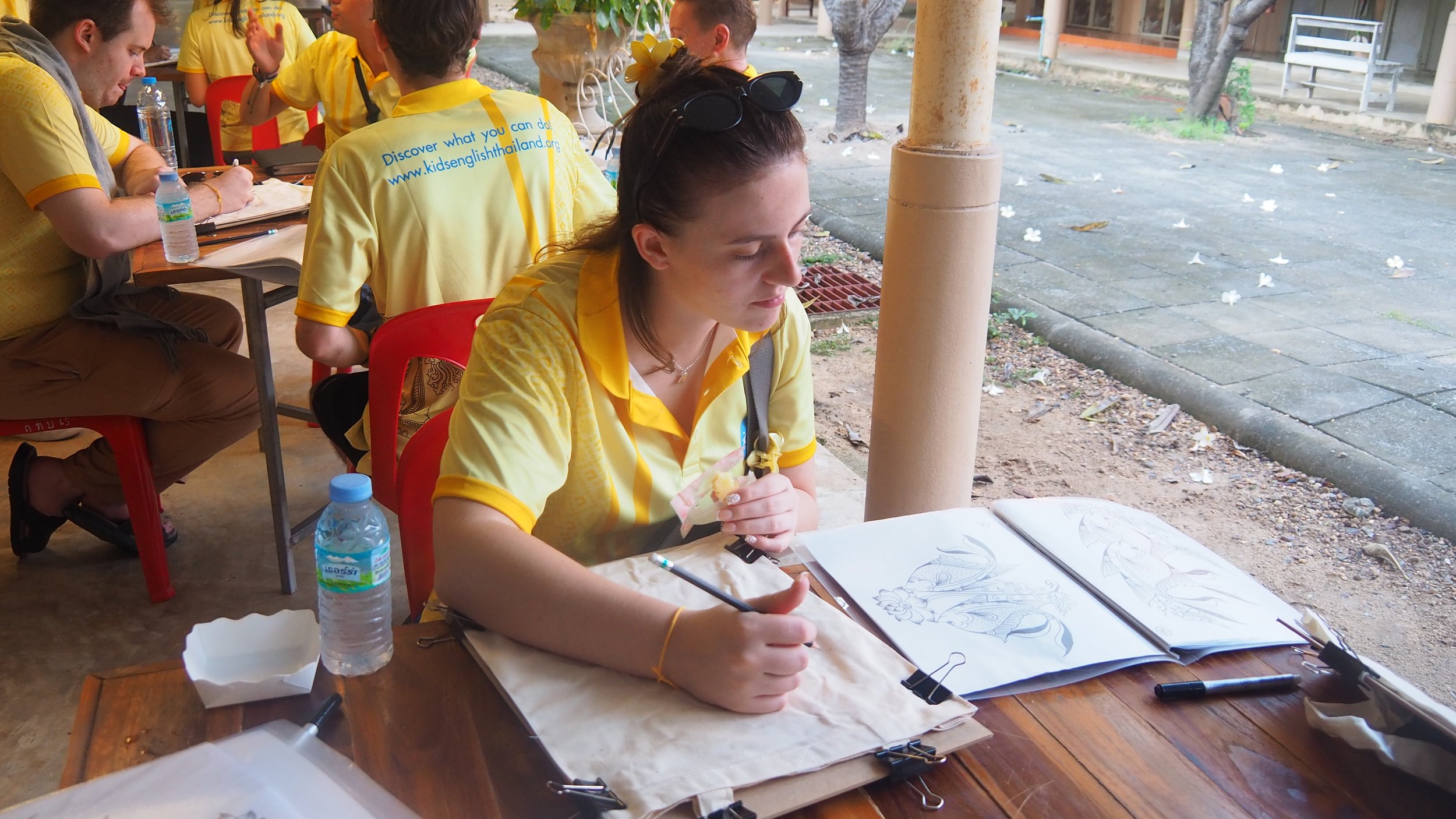 KET Teacher  in a yellow shirt with sunglasses on her head sits at a table outdoors, sketching in a drawing book with open pages displaying sketches of fish. 
