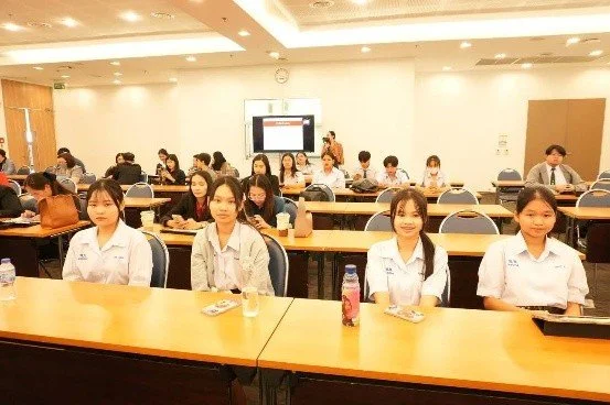 Kids English Thailand Image-A classroom with students seated at desks, facing forward, with a teacher at the front and a presentation screen behind her.