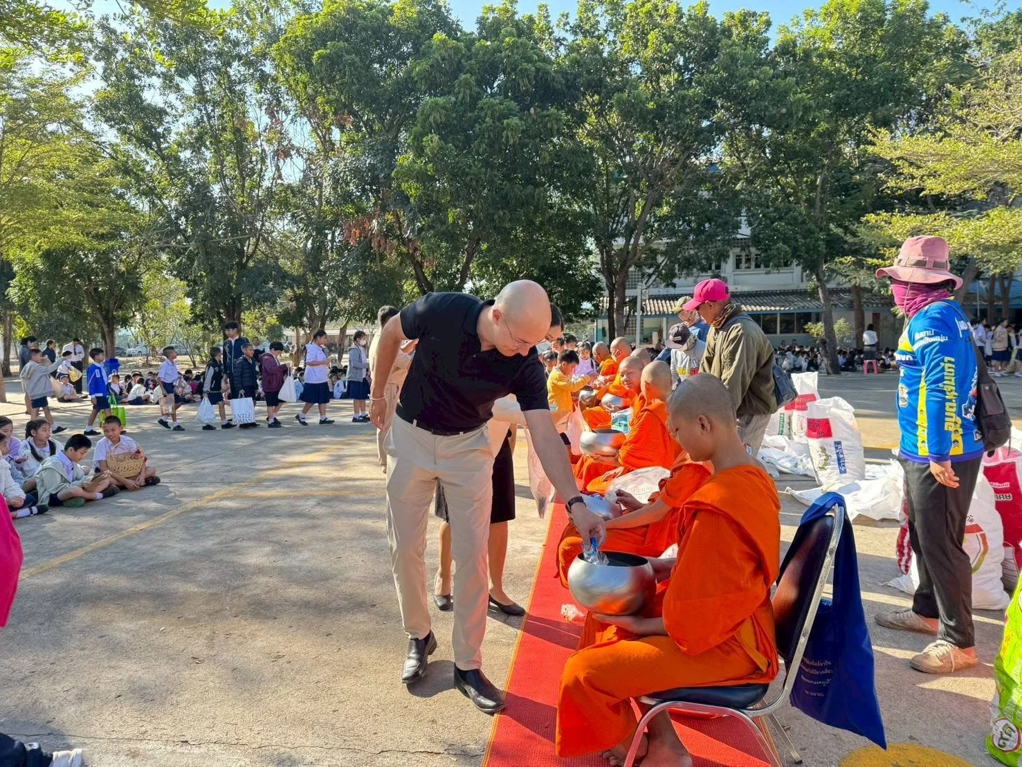 A man from Kids English Thailand offering alms to Buddhist monks dressed in orange robes seated on chairs outdoors, with children and adults in the background.
