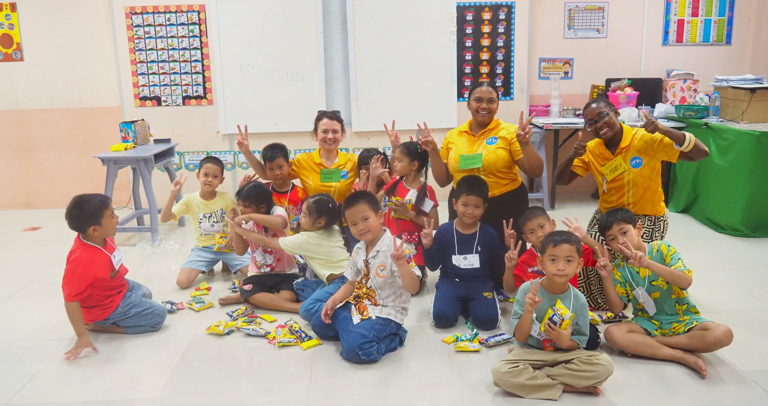 Group of Kids English Thailand children and three Kids English Thailand teachers posing happily inside a classroom, with children sitting on the floor, some holding snacks, and everyone making peace signs.
