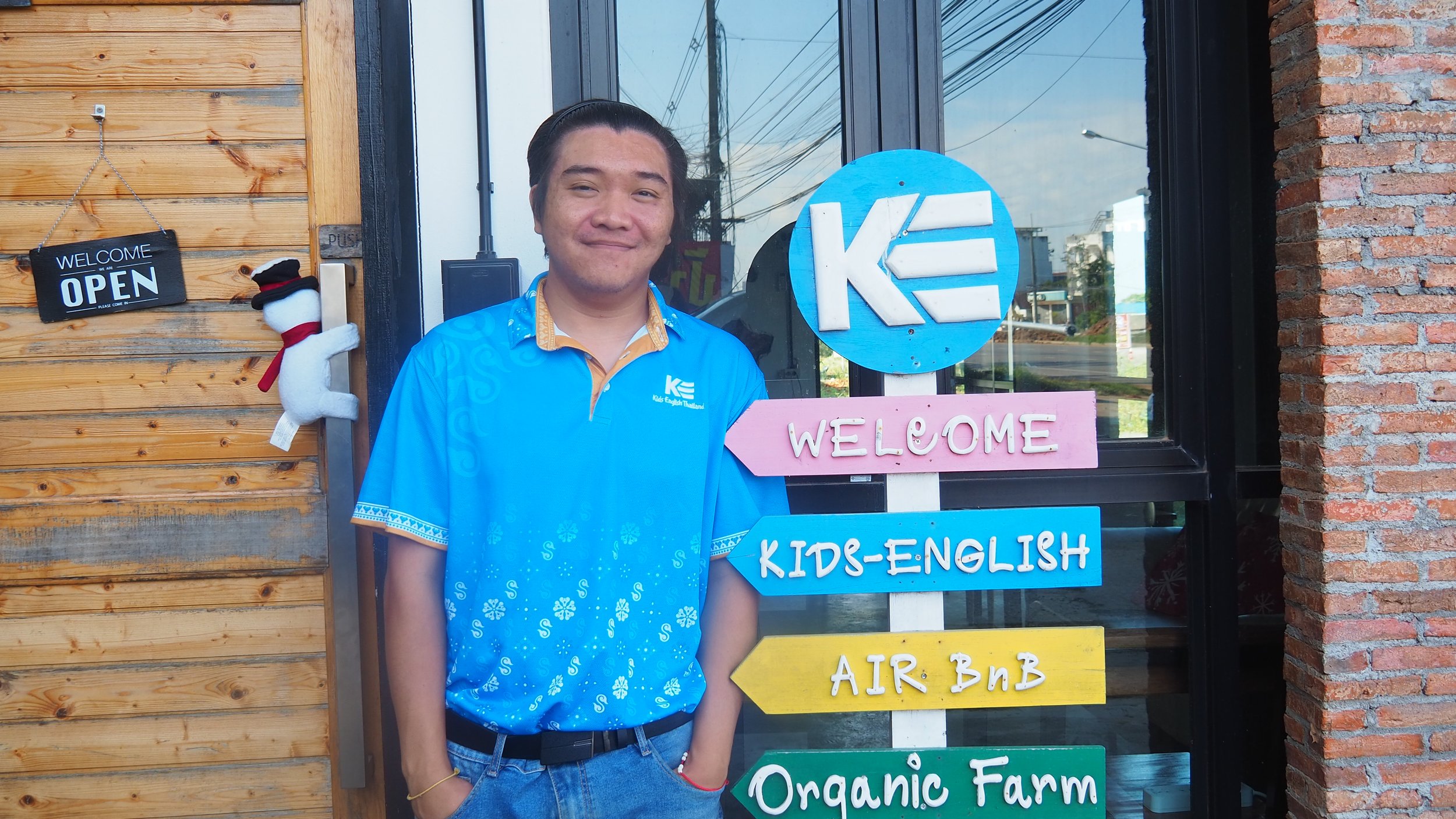 A young man standing outside a building with a wooden wall, smiling in front of colorful signs that read 'WELCOME,' 'KIDS-ENGLISH,' and 'AIR BNB.'