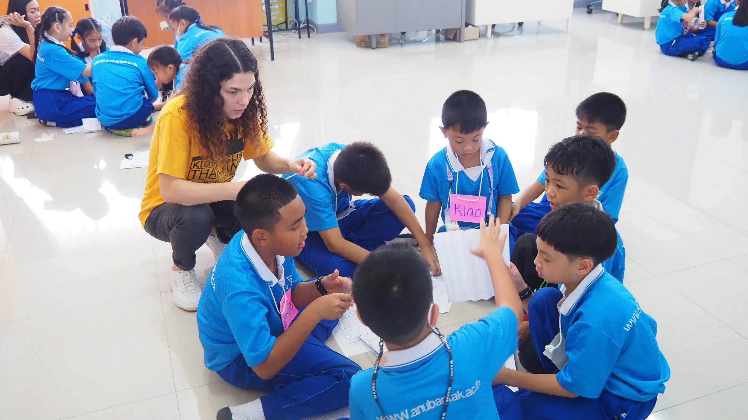 A group of young Asian students in blue uniforms sitting on the floor, engaging in a group activity with a female Kids English Thailand teacher in a yellow KET shirt supervising them.