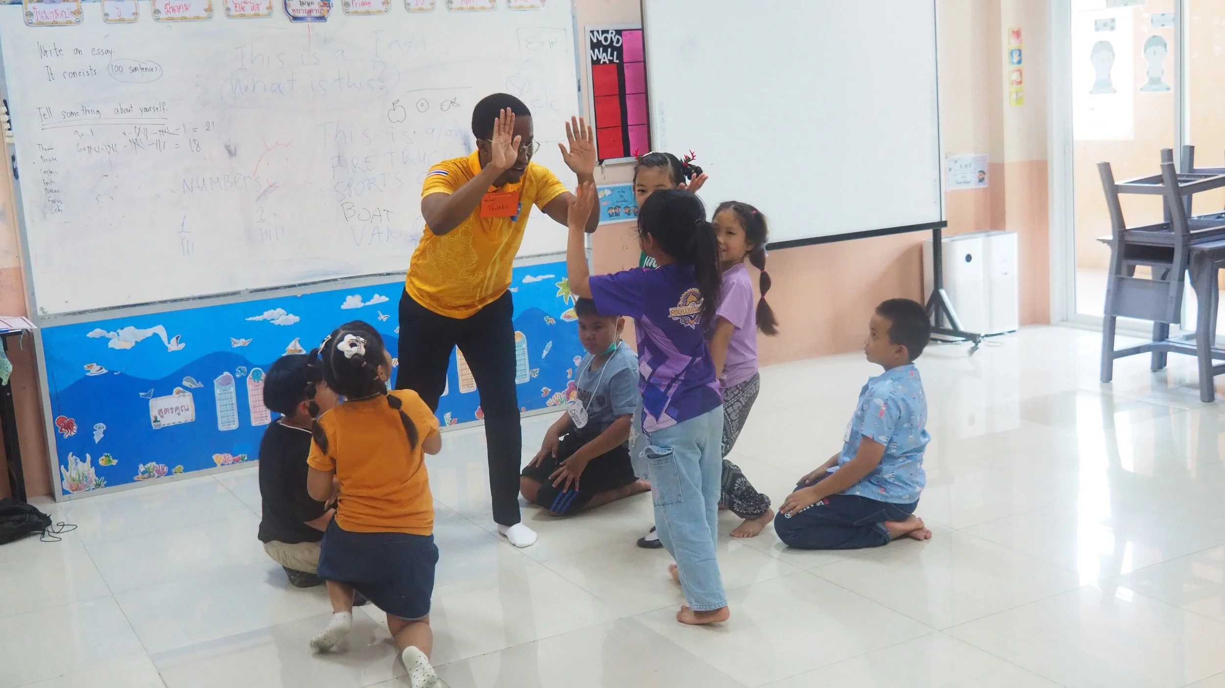 A  Kids English Thailand teacher giving high-fives to a group of children in a classroom. The children are kneeling or standing in front of a whiteboard, with some children wearing colorful clothes.