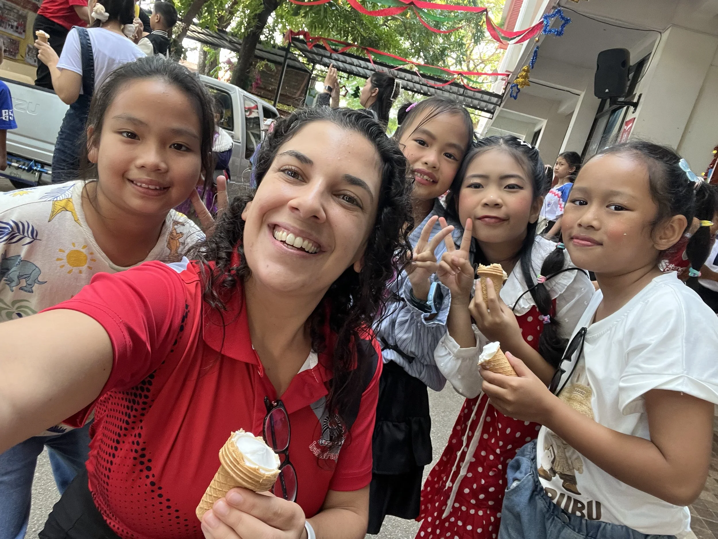 Group of smiling children and a Kids English Thailand teacher taking a selfie at an outdoor celebration, with ice cream cones in hand, decorations hanging above, and people in the background.