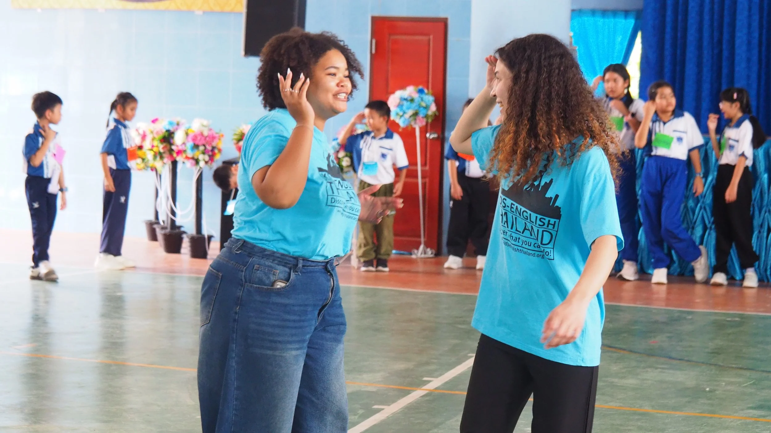 Two Kids English Thailand teachers engaging in a dance or game in a school gymnasium, with children standing in line and watching in the background.