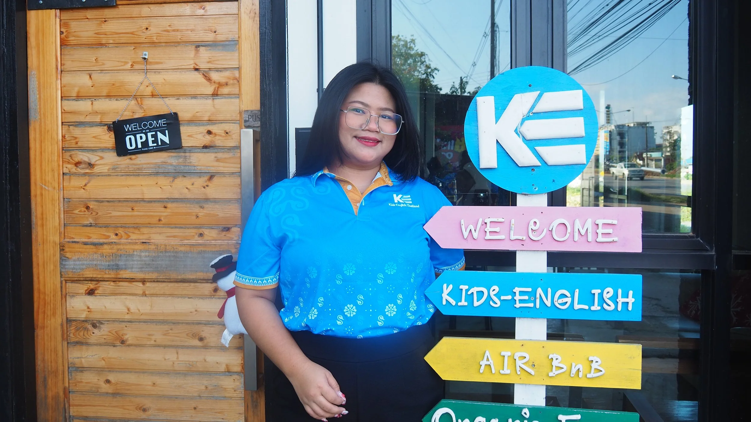 A woman with long black hair, glasses, and red flowers in her hair, smiling and standing next to a colorful sign outside a building. The sign reads 'WELCOME' in pink letters, with blue arrows pointing to 'KIDS-ENGLISH' and another direction.