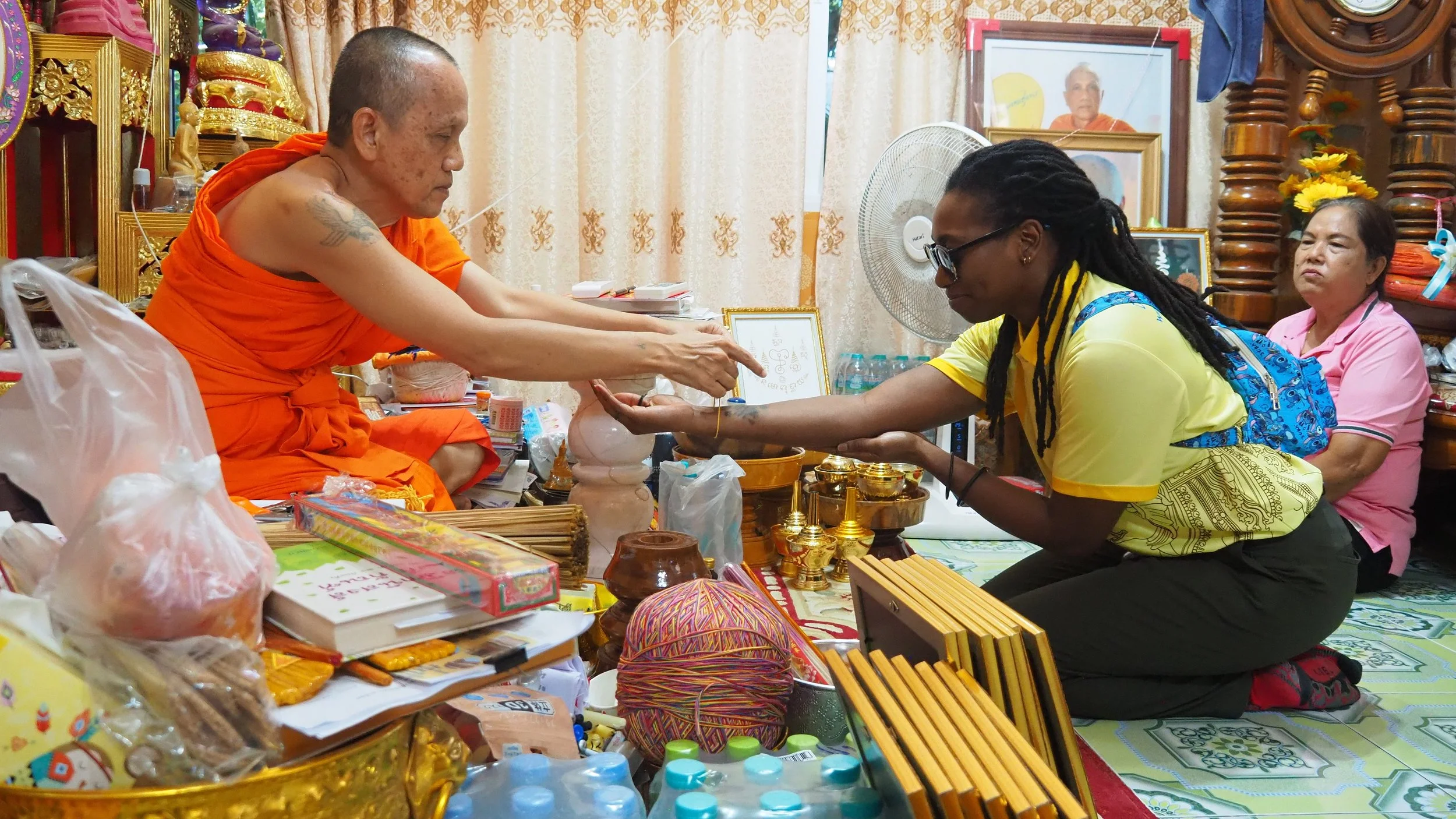 A Kids English Thailand teacher in a yellow shirt and black pants kneels on the floor, receiving a blessing or ritual from a Buddhist monk dressed in orange robes, who is seated among offerings and religious items. 