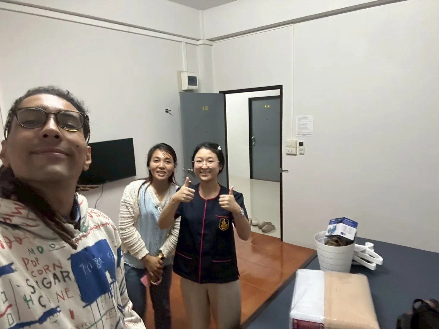 Three people from Kids English Thailand standing indoors, smiling and giving thumbs up, with a table and a door in the background.