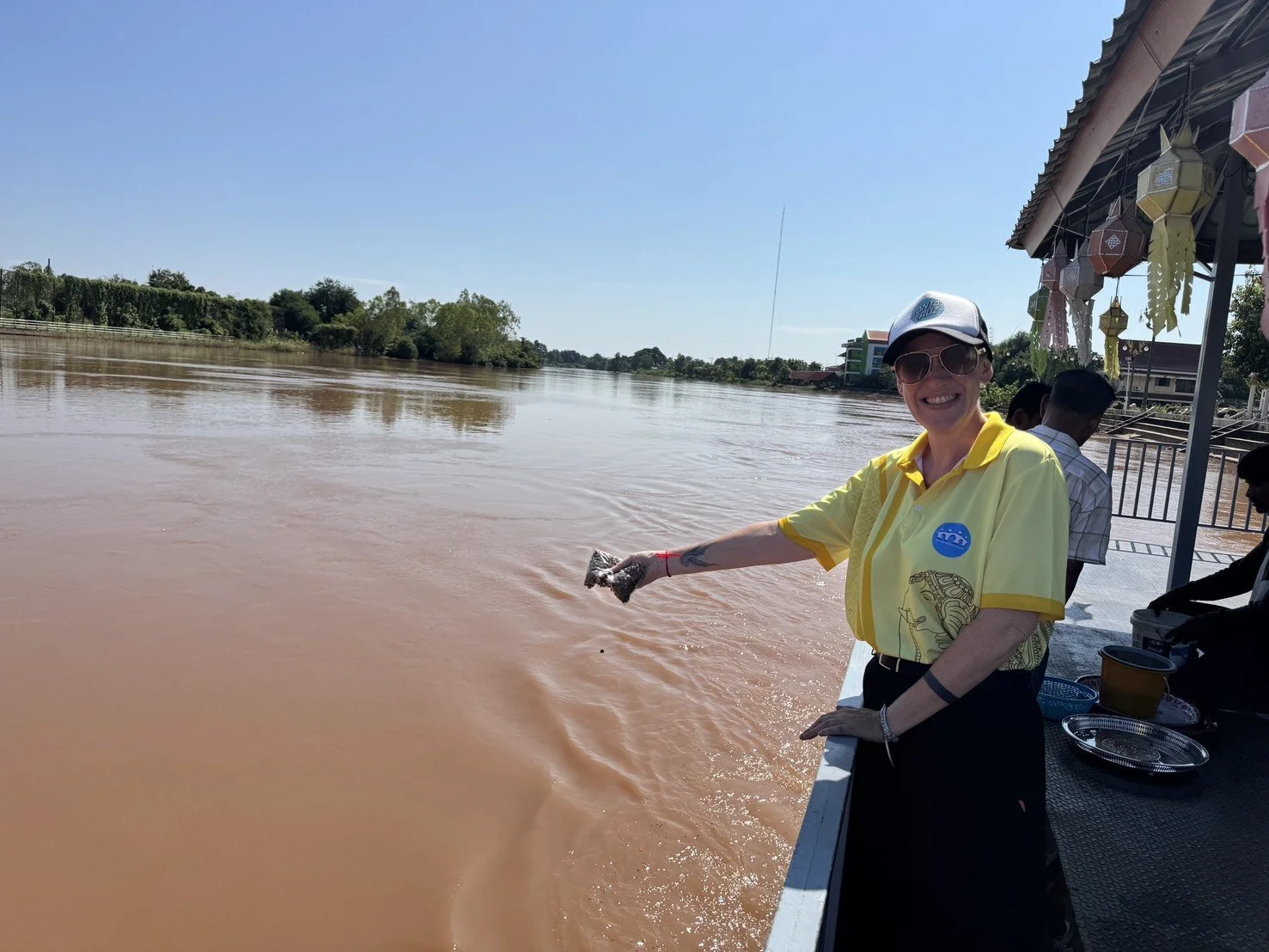 A smiling Kids English Thailand teacher in a yellow KET shirt with turtle design, sunglasses, and a white cap, throwing an object into a river from a boat on a sunny day.