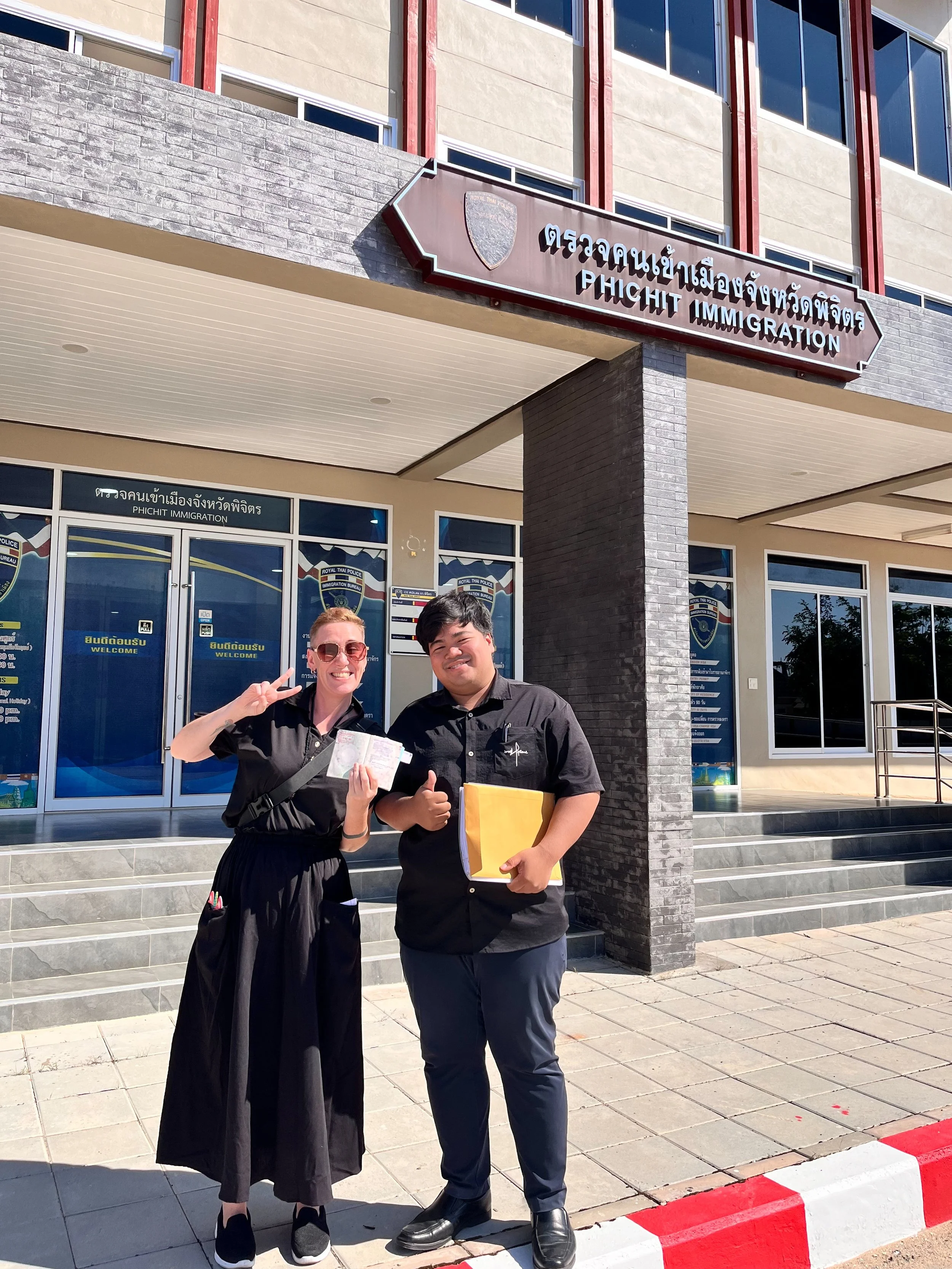 Two Kids English Thailand teachers standing in front of a building labeled 'Phichit Immigration' with one holding a passport and the other holding folders and making a peace sign.