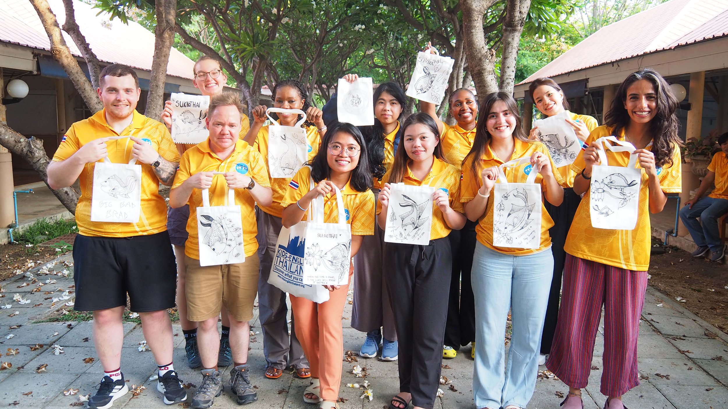 A group of diverse young Kids English Thailand teachers wearing yellow shirts outdoors, holding up drawings and tote bags featuring fish illustrations, smiling at the camera.