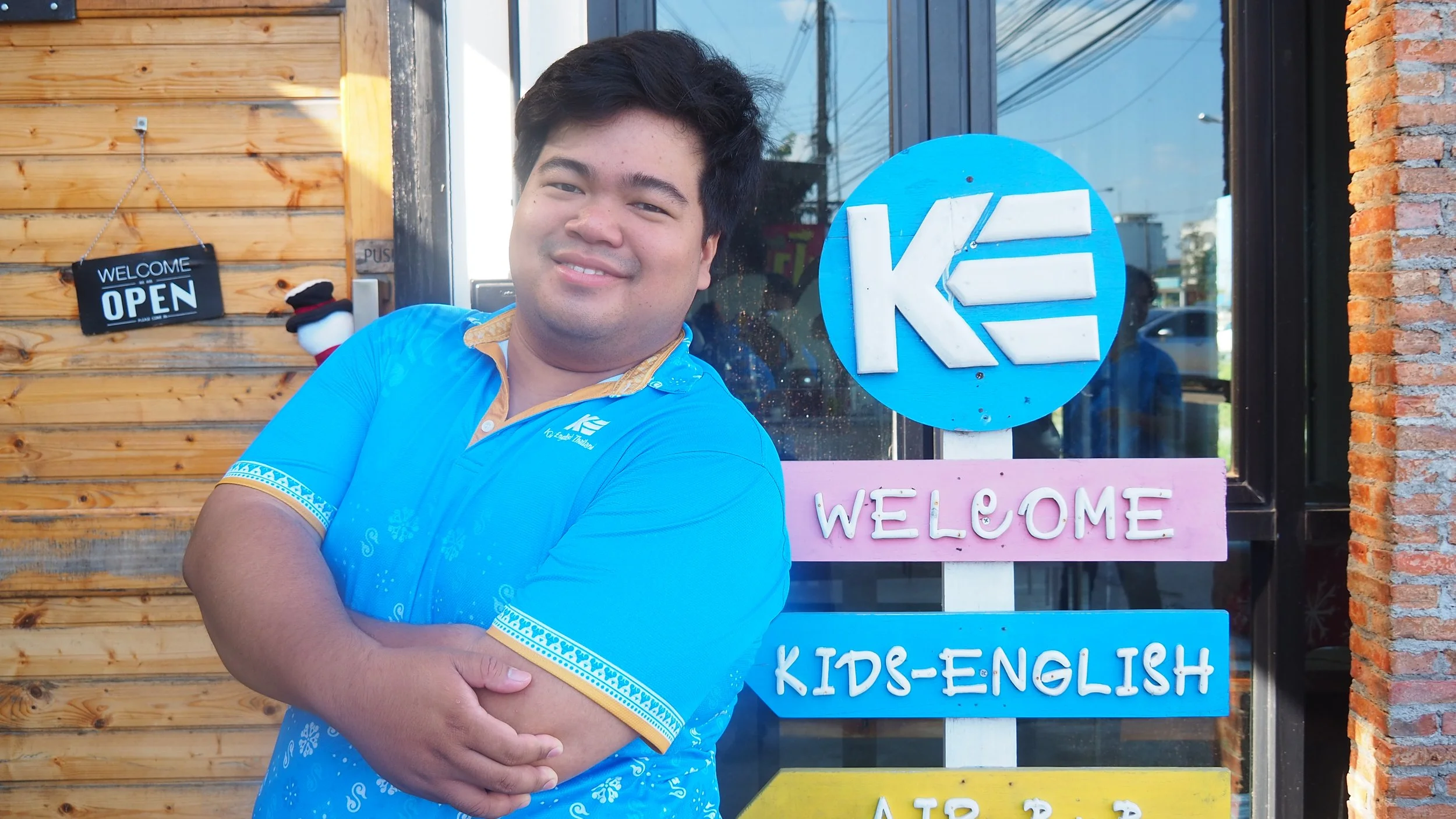 A young man with black hair, smiling, wearing a bright blue shirt, standing in front of a store with a wooden and brick wall, and colorful signs that read "WELCOME," "KIDS-ENGLISH," and others. A sign on the door says "WELCOME OPEN."