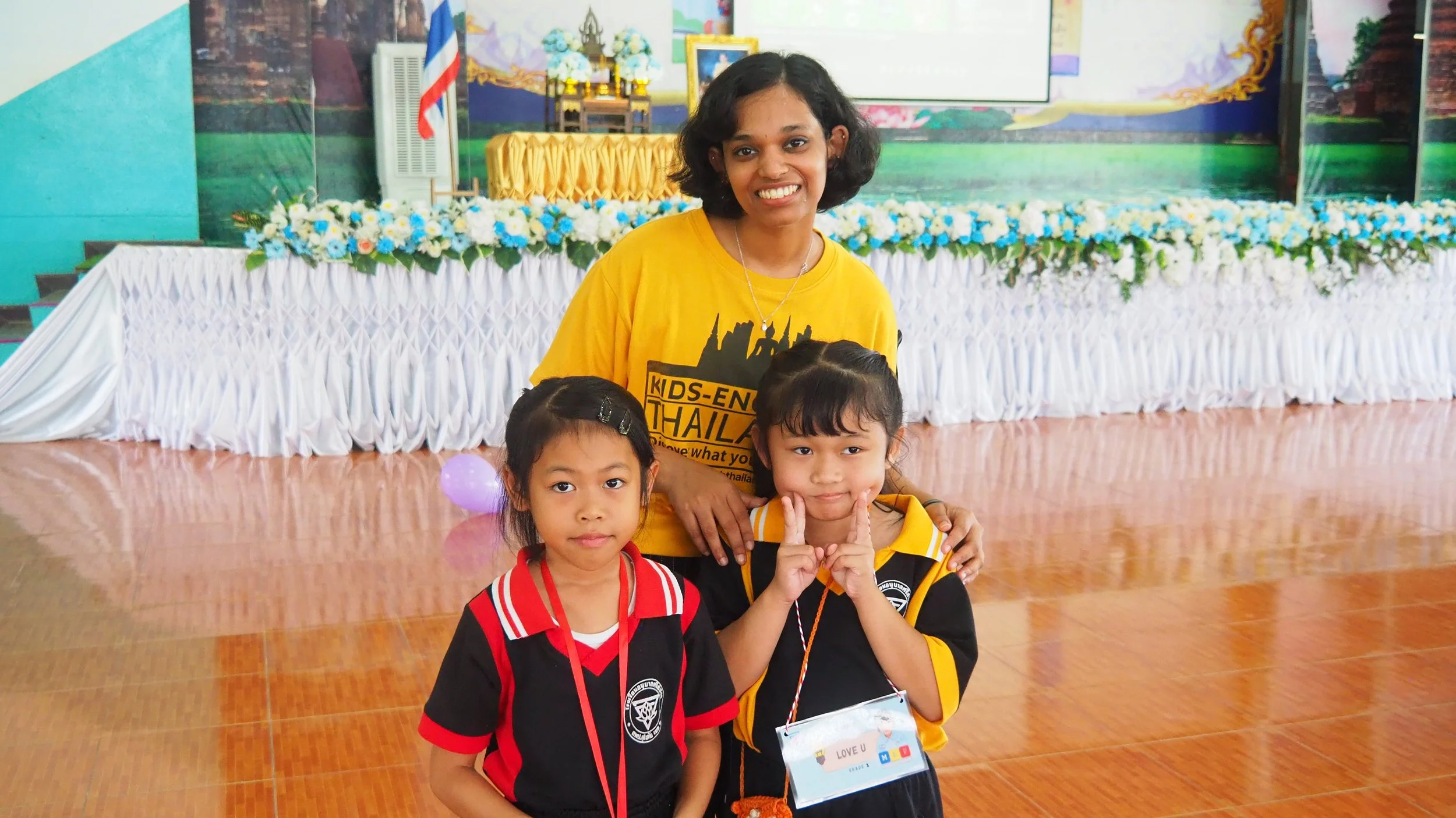 A Kids English Thailand teacher and two young girls smiling in a decorated indoor space. The woman is standing behind the girls, wearing a yellow T-shirt, and the girls are wearing black and red school uniforms. T