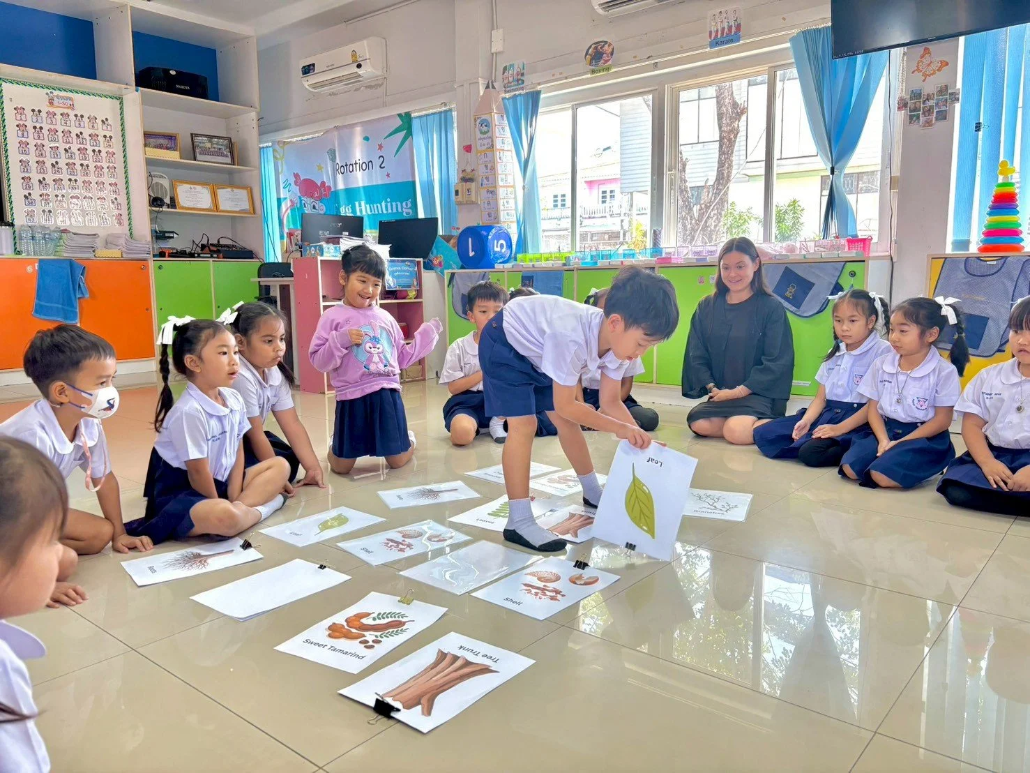 Kids sitting on the floor in a classroom, watching a boy in the center arrange large pictures of leaves and trees, with a teacher from Kids English Thailand observing. The classroom is colorful with educational posters and large windows.