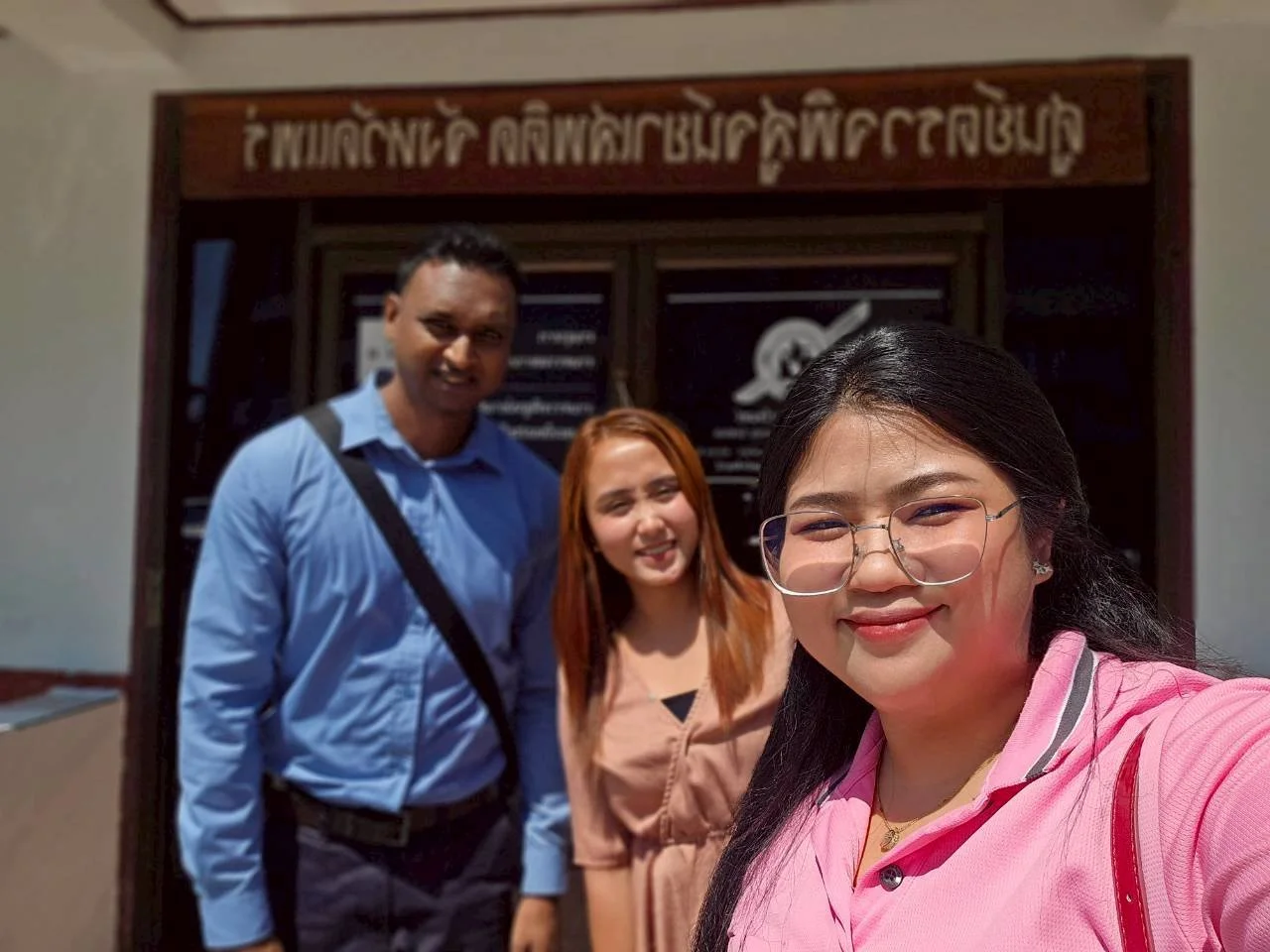 Three people from Kids English Thailand taking a selfie in front of a building with a sign written in a non-Latin script. The person in the foreground is a woman with glasses, wearing a pink shirt. 