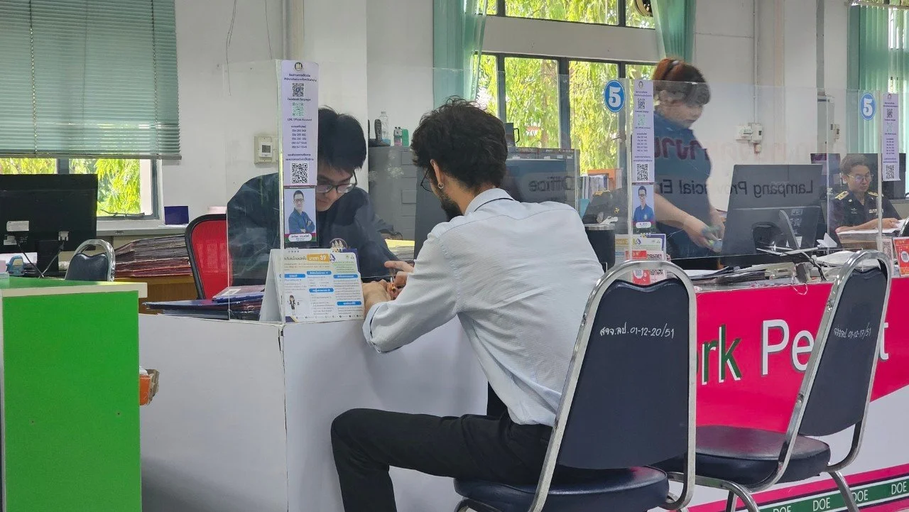 A man from Kids English Thailand in a white shirt sitting at a counter talking to a person from Kids English Thailand in glasses behind a plastic barrier, with other people working in the background at an office or bank.