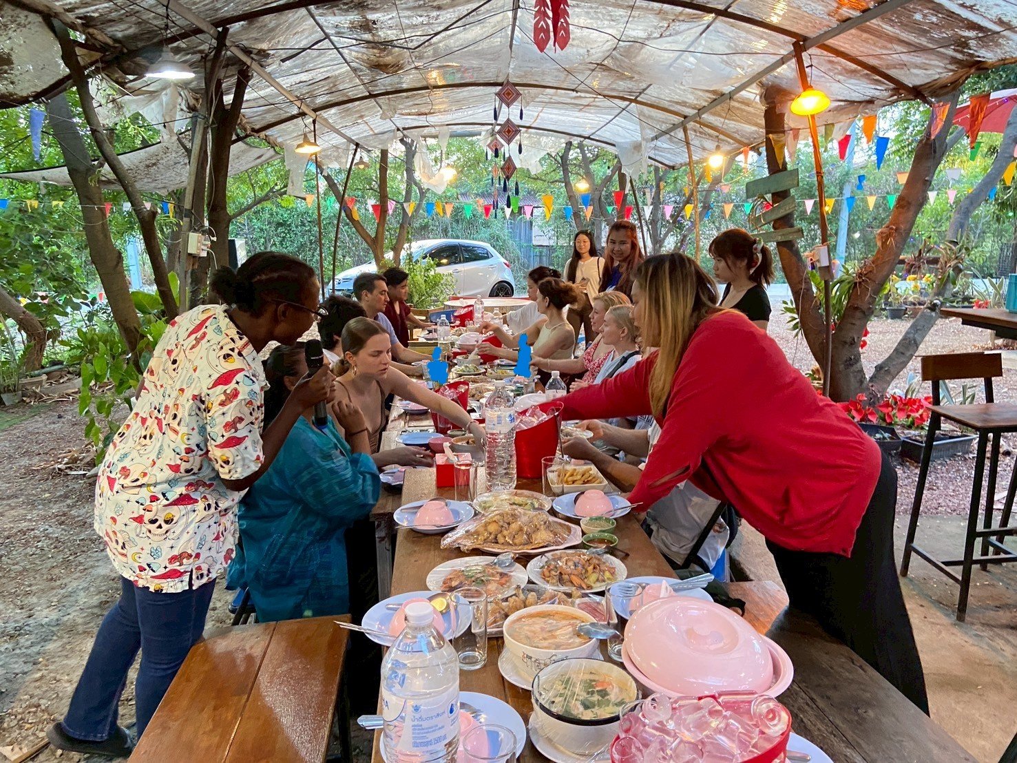 People gathered around a long outdoor table under a makeshift canopy with colorful bunting, sharing a meal with various dishes and drinks in a garden setting. Kids English Thailand experience!