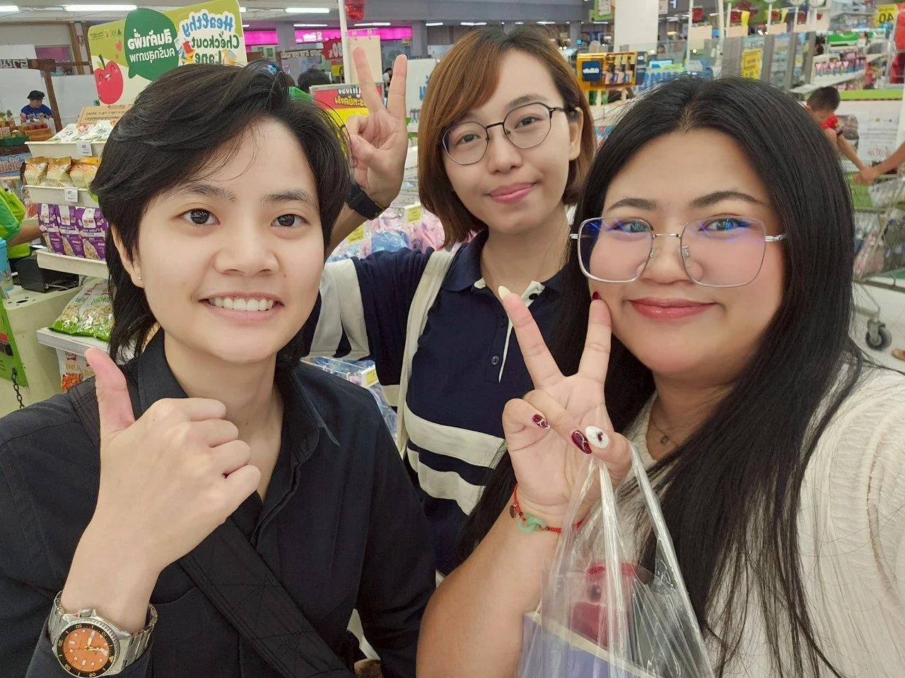 Three women from Kids English Thailand smiling and making peace signs in a grocery store, with shelves of snacks and products in the background.