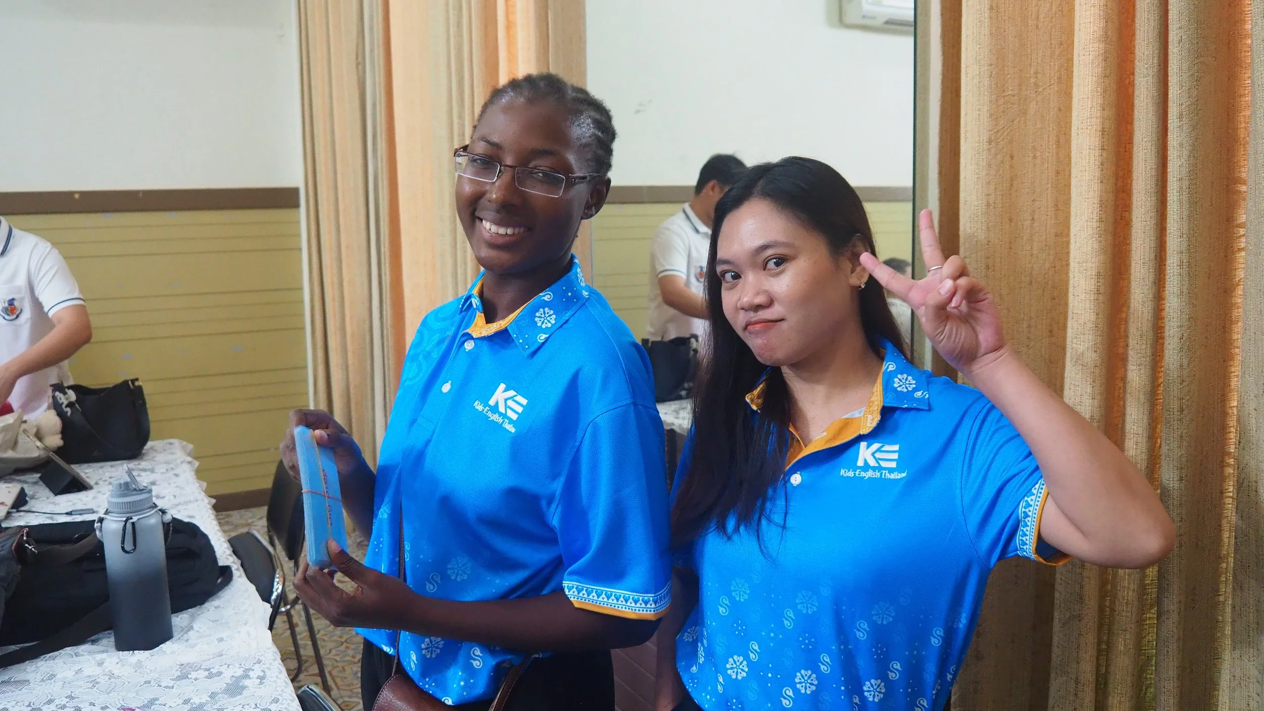 Two young Kids English Thailand teacher wearing blue KET shirts with Kids English Thailand logo, standing indoors, smiling; one making a peace sign gesture.