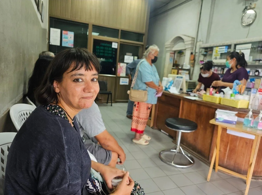 A woman from Kids English Thailand sitting in a waiting area, with a pharmacy counter in the background, where three staff members and a customer are standing, wearing face masks.